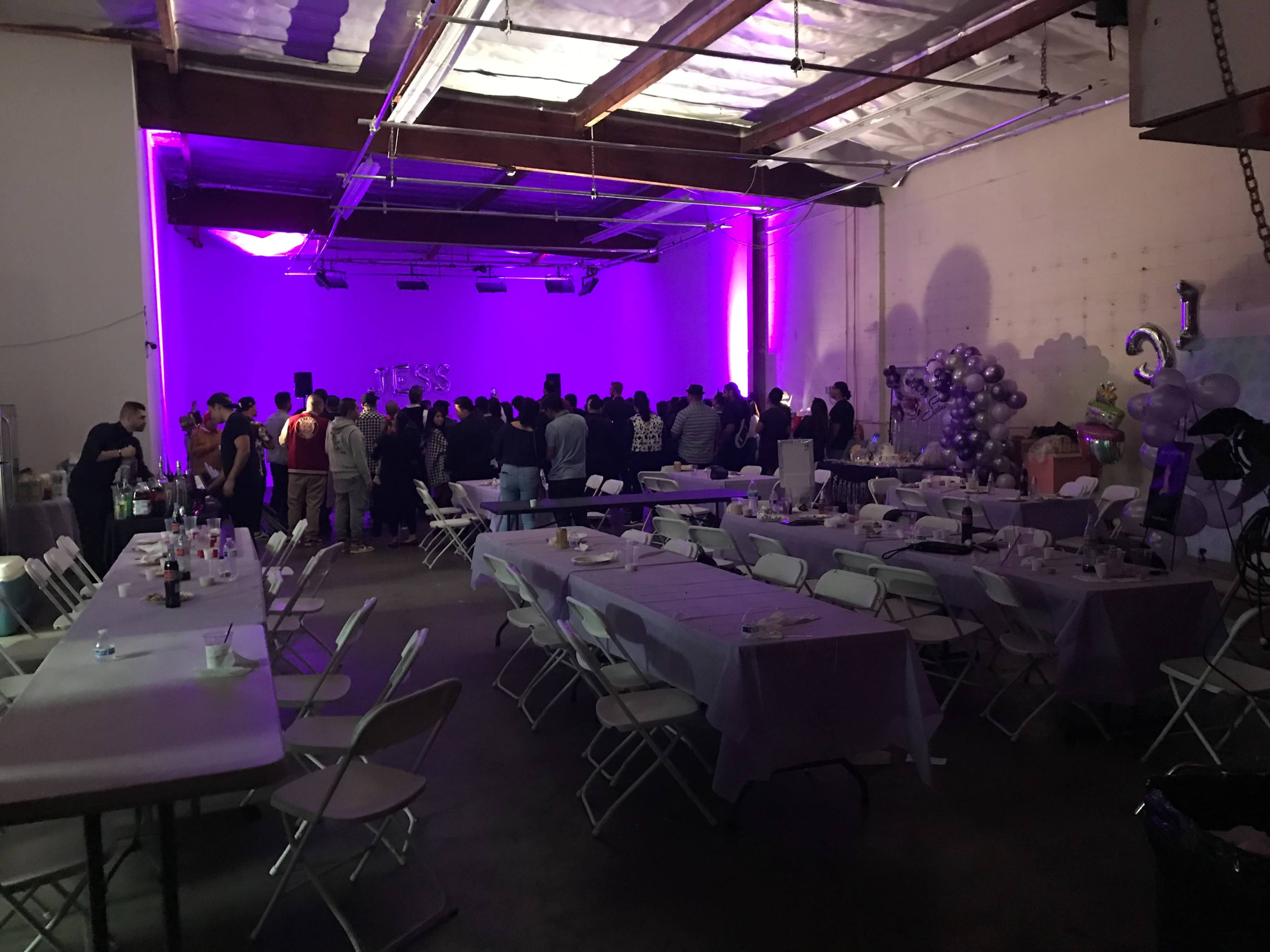 A large indoor space is filled with people gathered in front of a stage, while tables and chairs are arranged for an event.