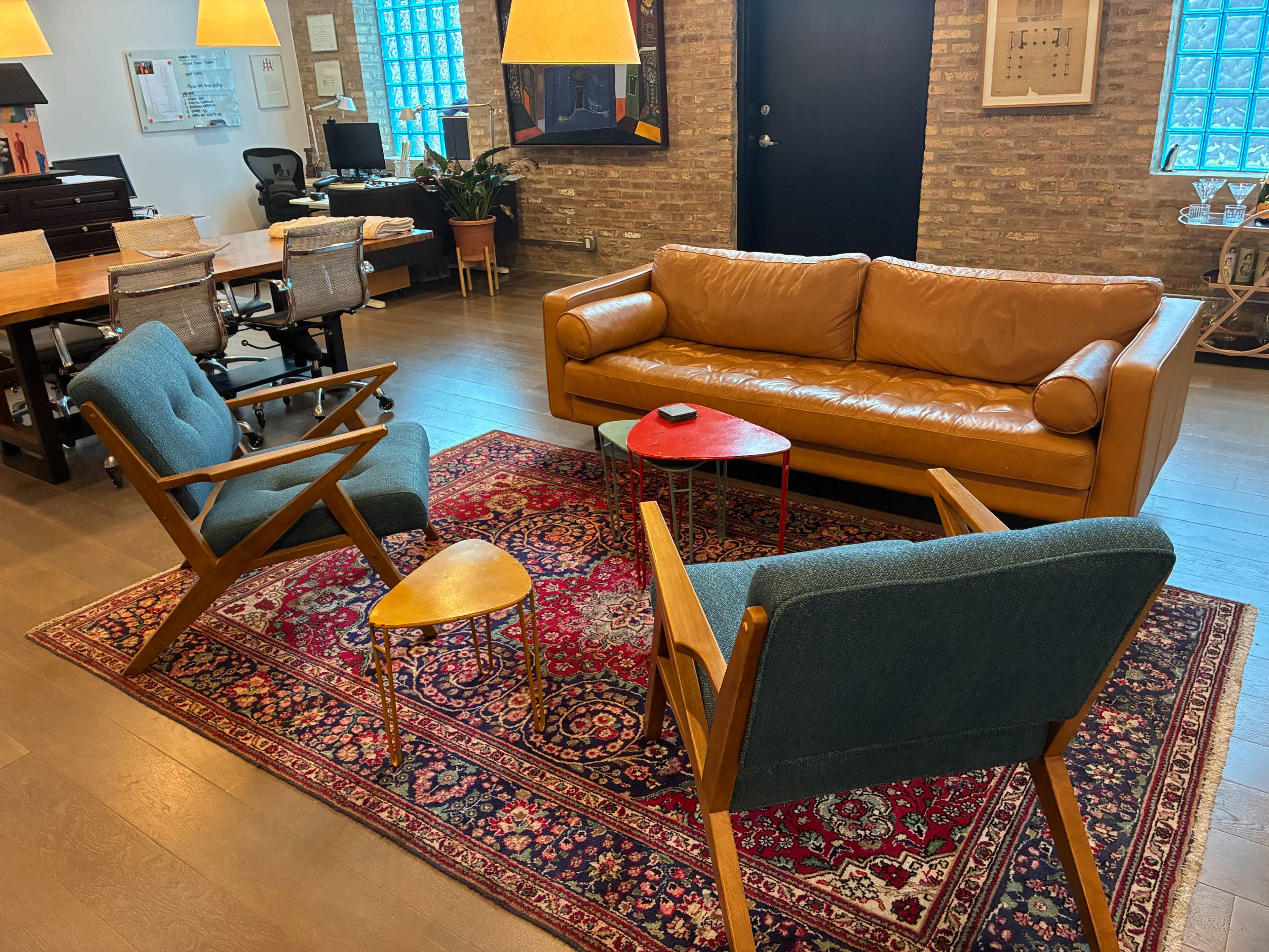 A modern office space features a brown leather sofa, two blue armchairs, a small wooden side table, and a patterned area rug, all arranged around a central seating area.