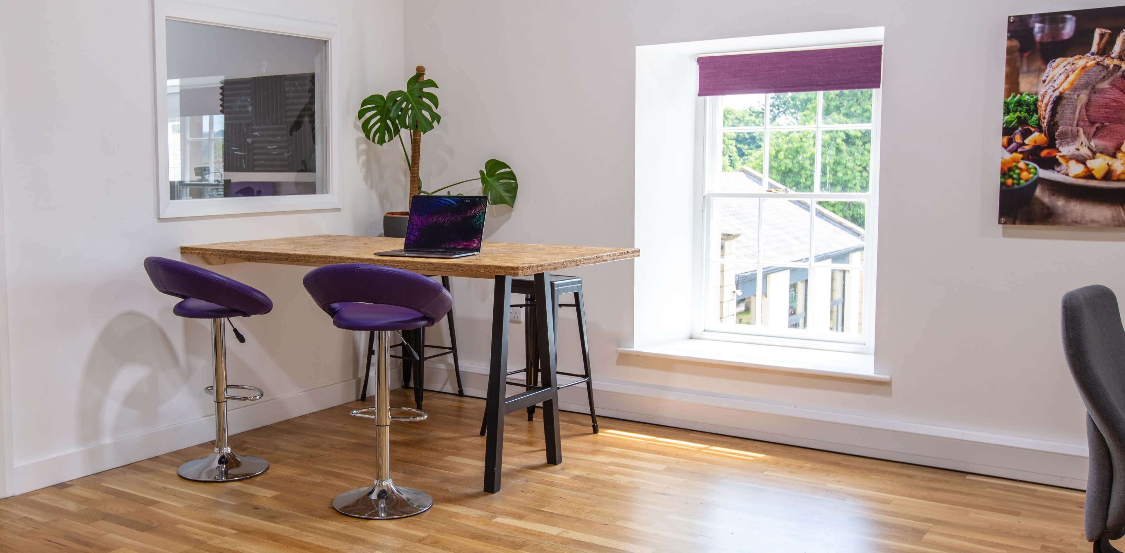 A modern workspace features a wooden table with a laptop, purple bar stools, and a large window overlooking a garden.