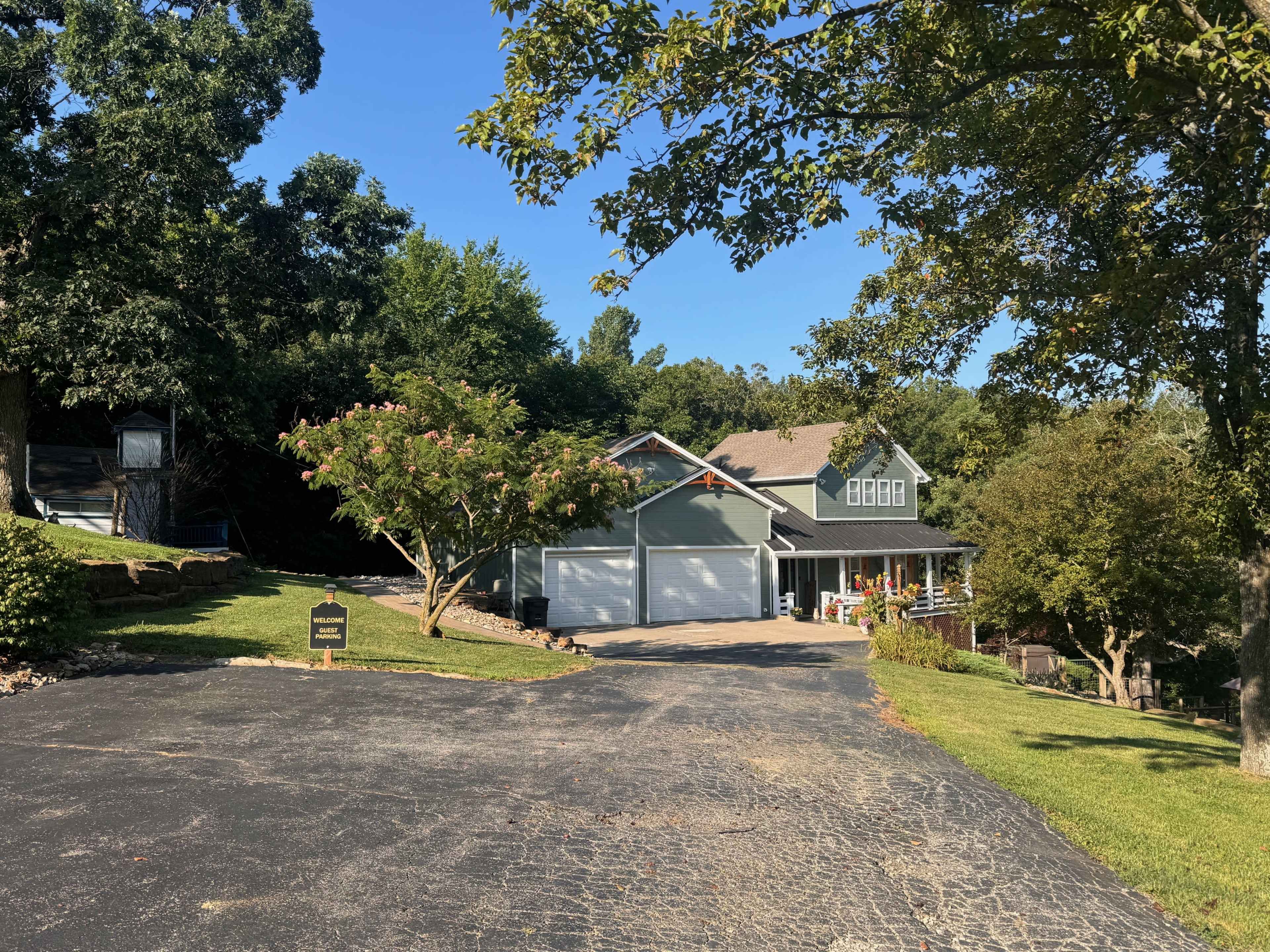A green house with a two-car garage sits at the end of a driveway surrounded by trees and lawn.