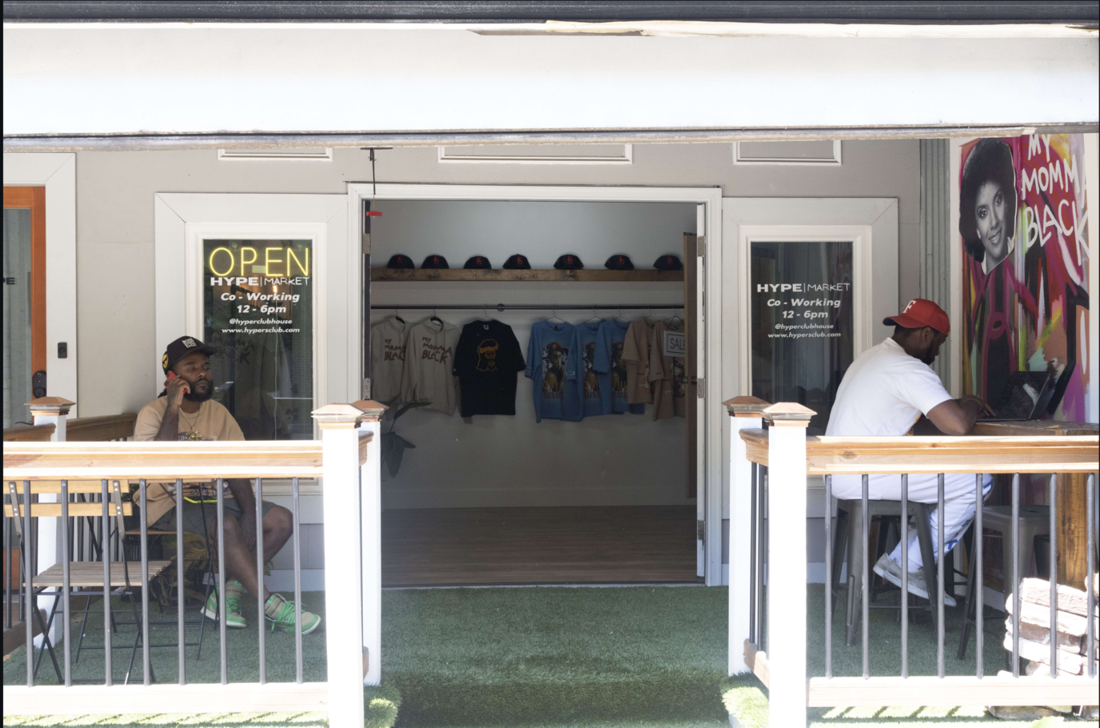 Two men sit at a table outside a storefront labeled "OPEN" and "HYPE MARKET," which displays clothing and artwork inside.