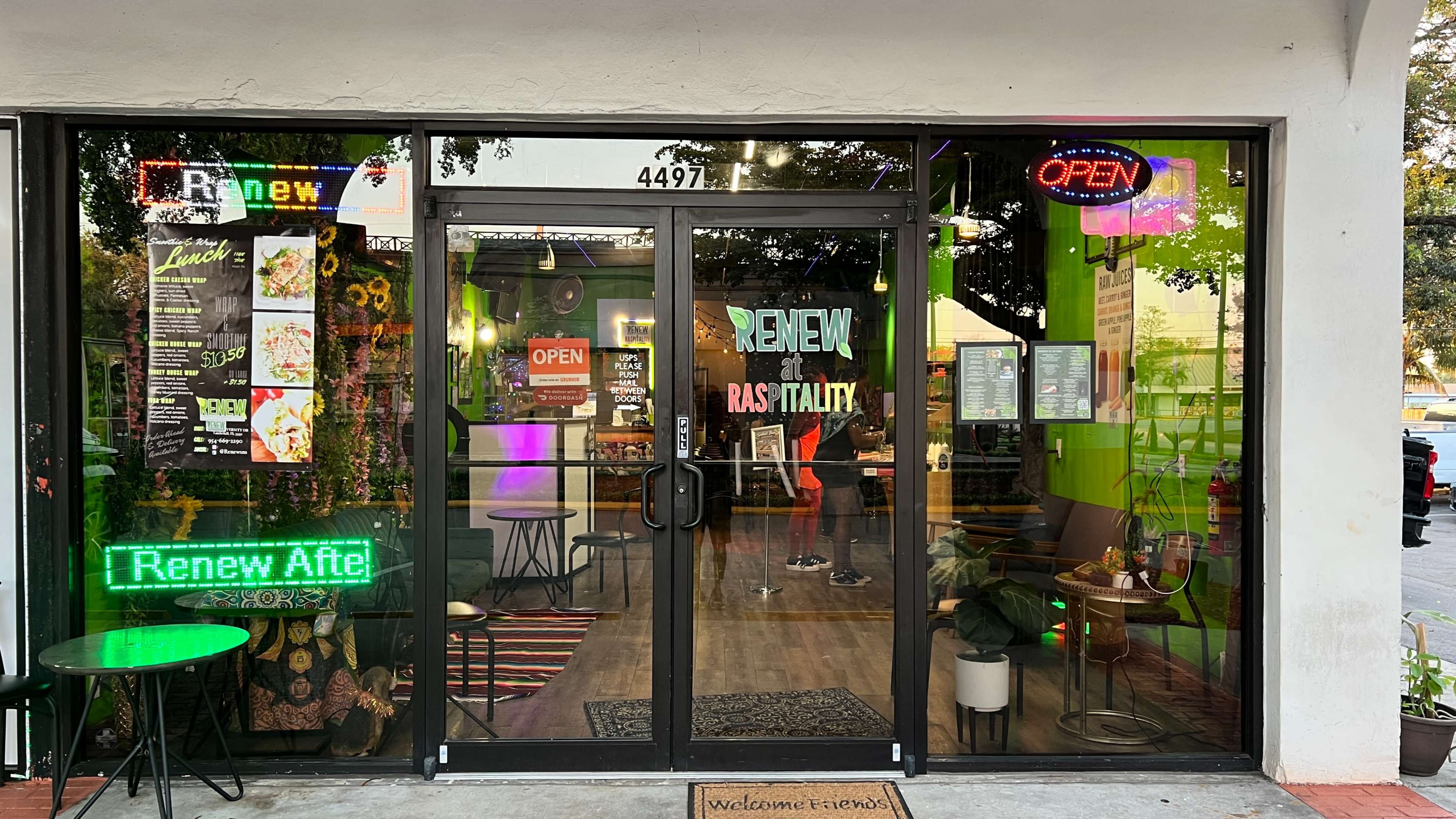 The image shows the entrance of a brightly lit cafe called "Renew" with large glass doors displaying various signs, including "OPEN" and "Renew After."