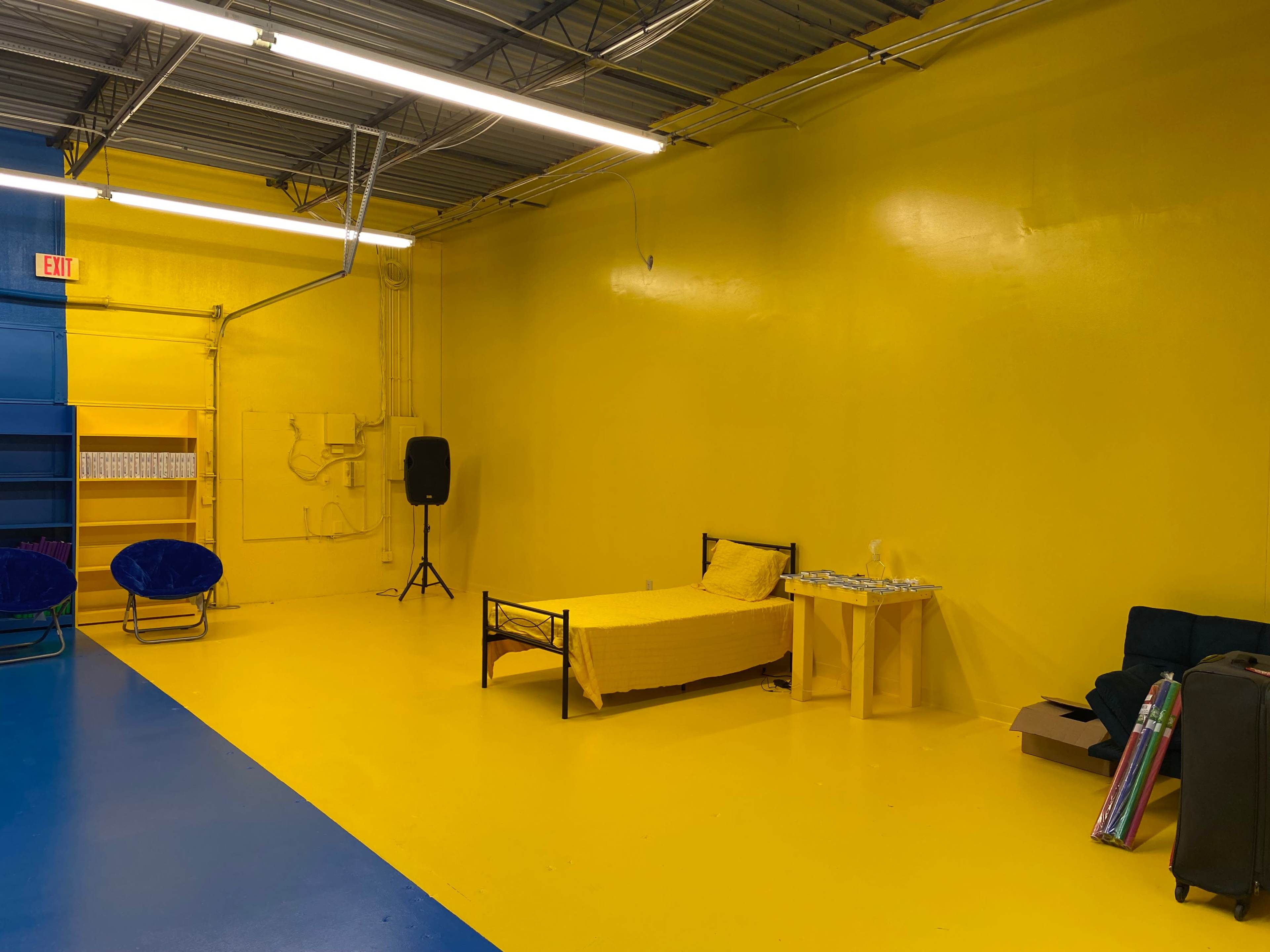 A room with yellow walls and flooring, featuring a bed with a yellow bedspread, a small table, and a blue area on one side.