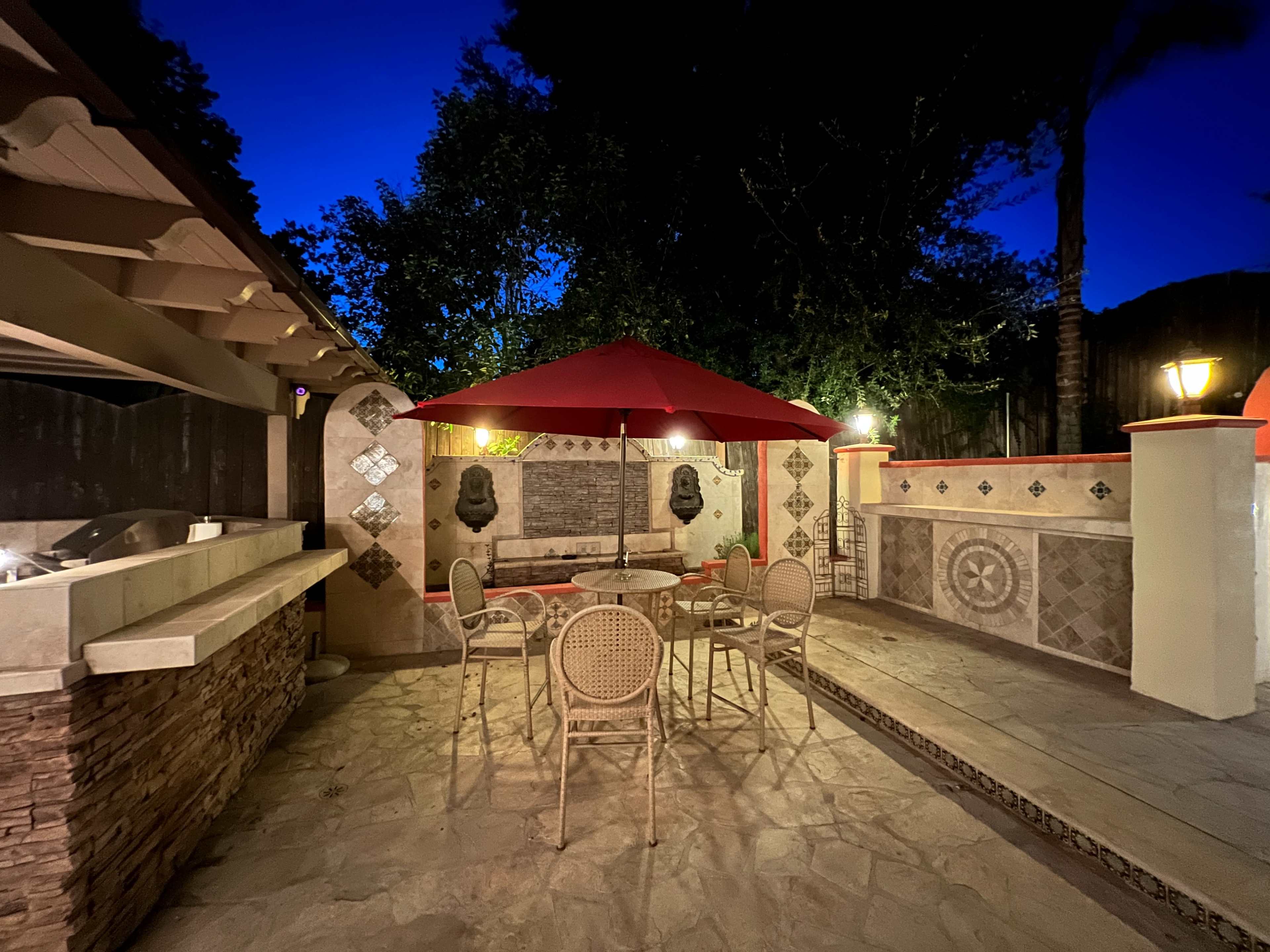 A patio area features a table and chairs under a red umbrella, with a stone wall and outdoor kitchen in the background.