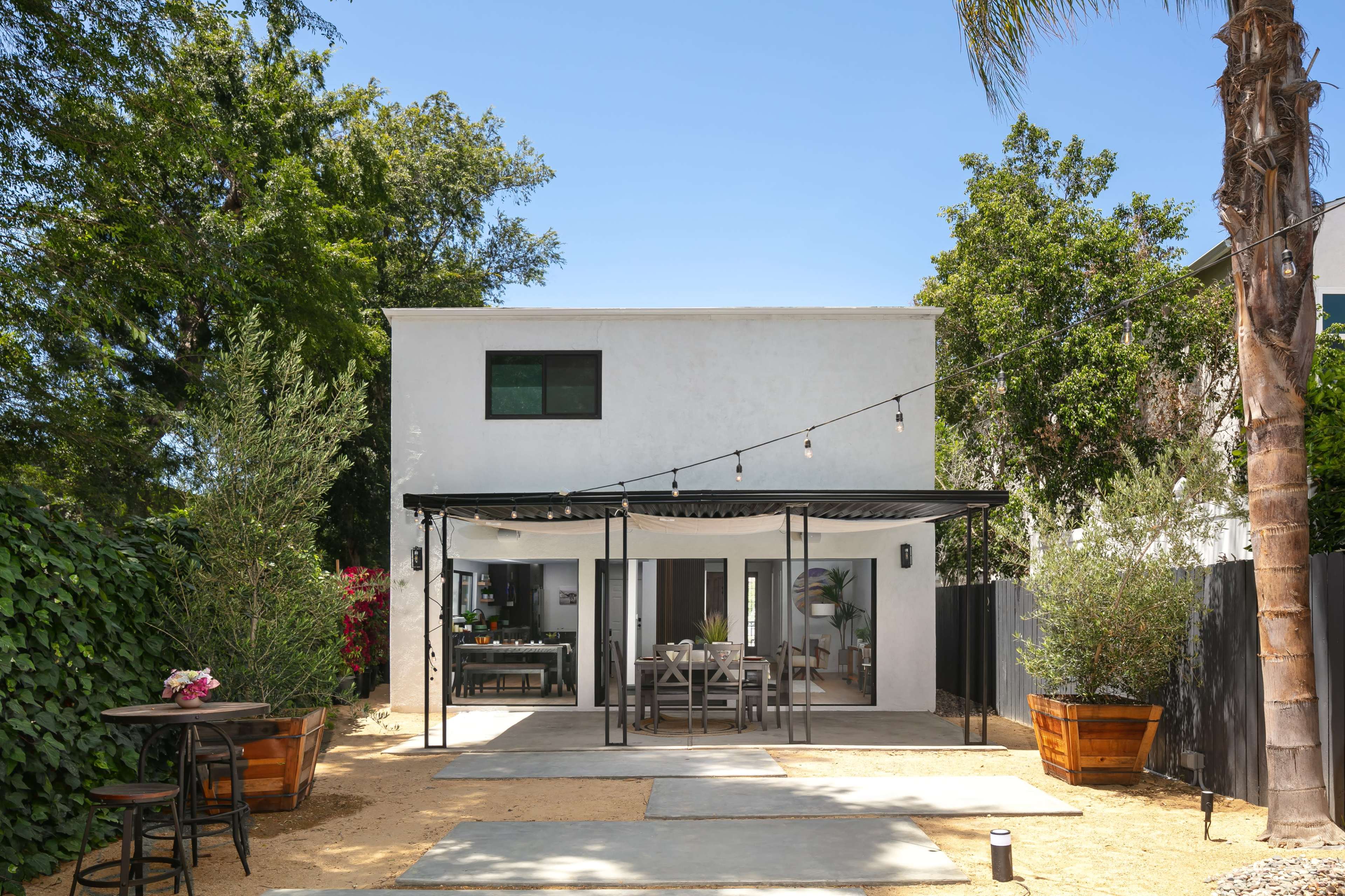 A two-story house with a covered patio area and outdoor dining set is surrounded by greenery and landscaping.