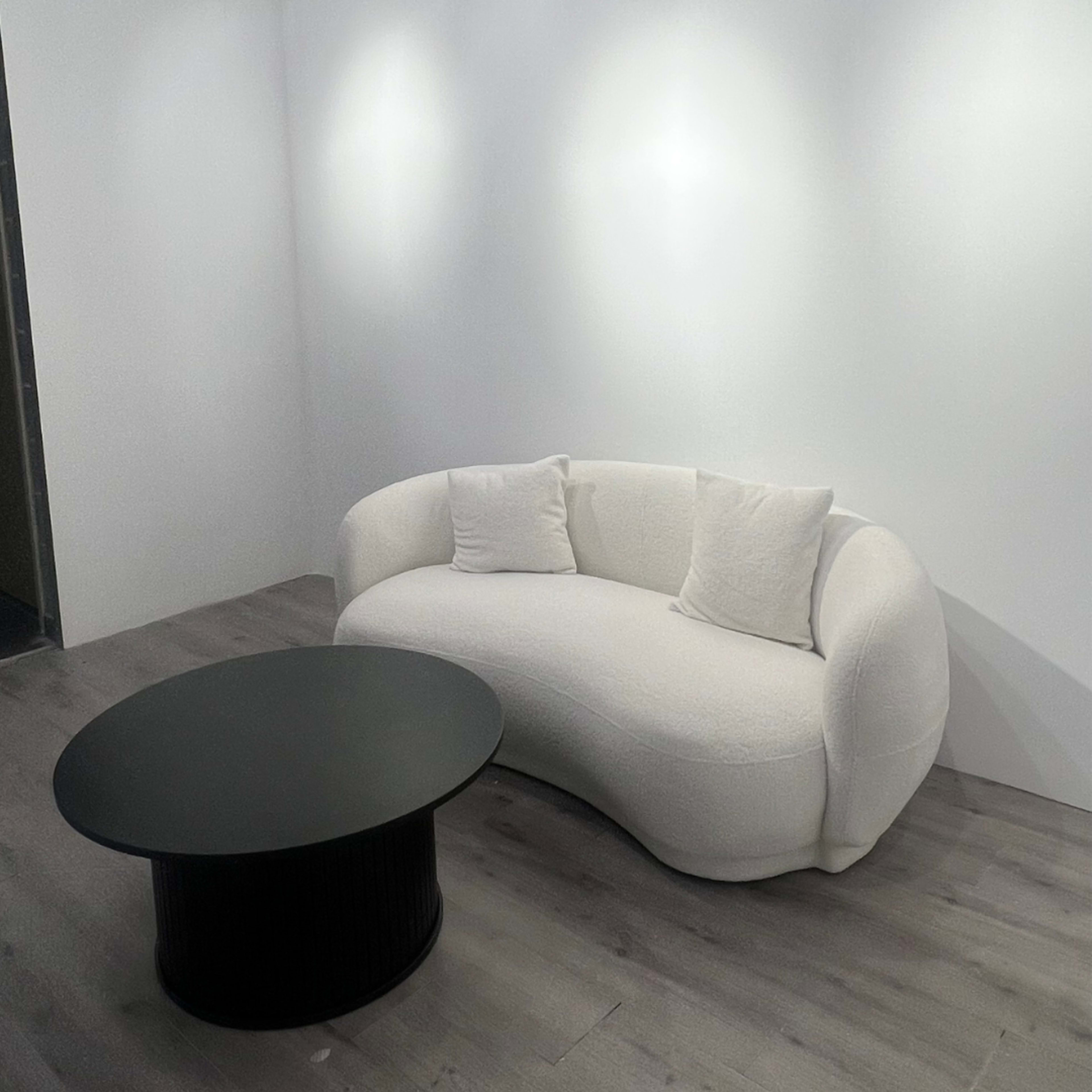 A white curved sofa with two pillows is positioned near a round black coffee table in a minimalist room with light-colored flooring.