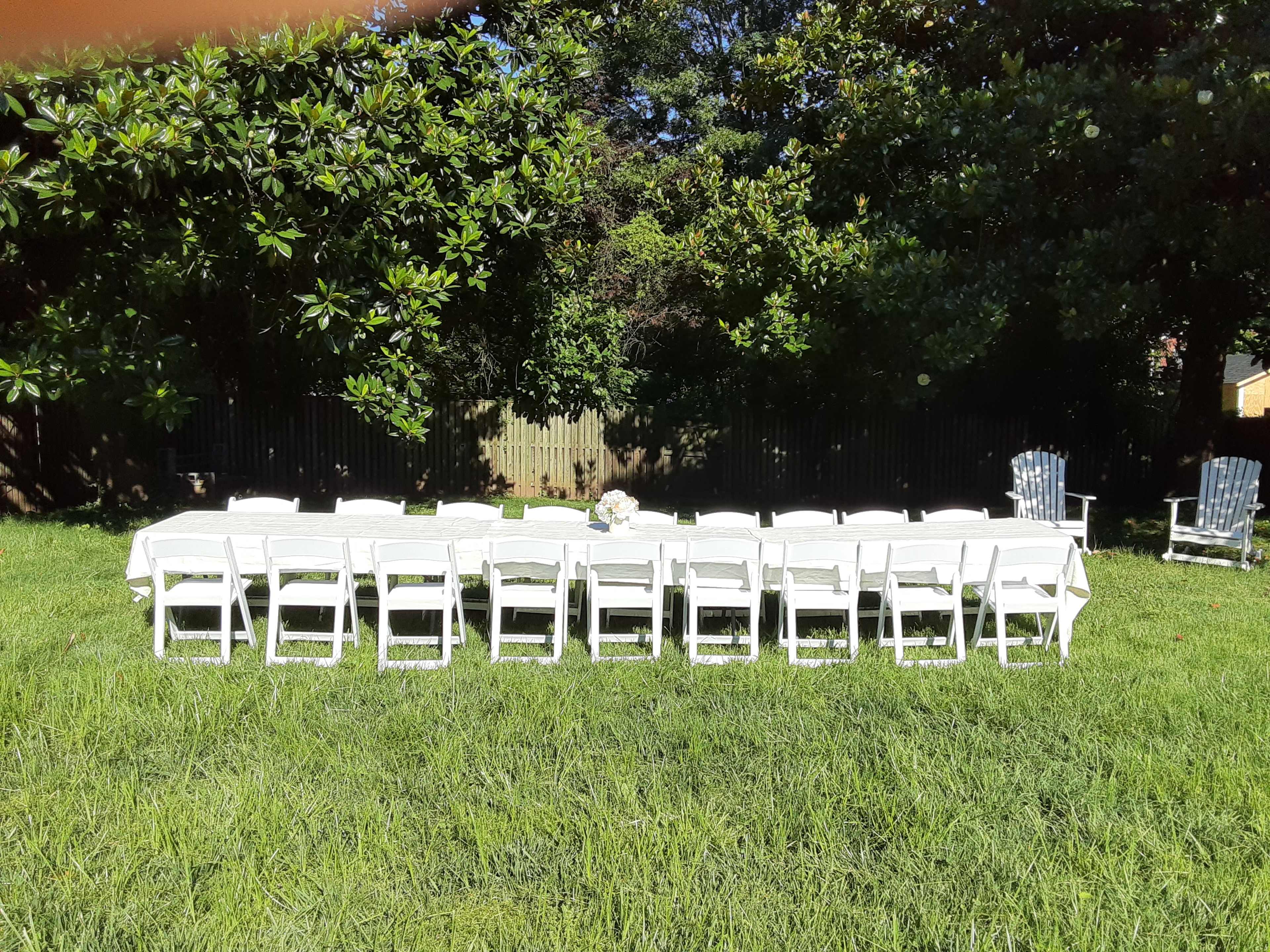 A long table with white tablecloths and chairs is set up on a grassy area surrounded by trees.