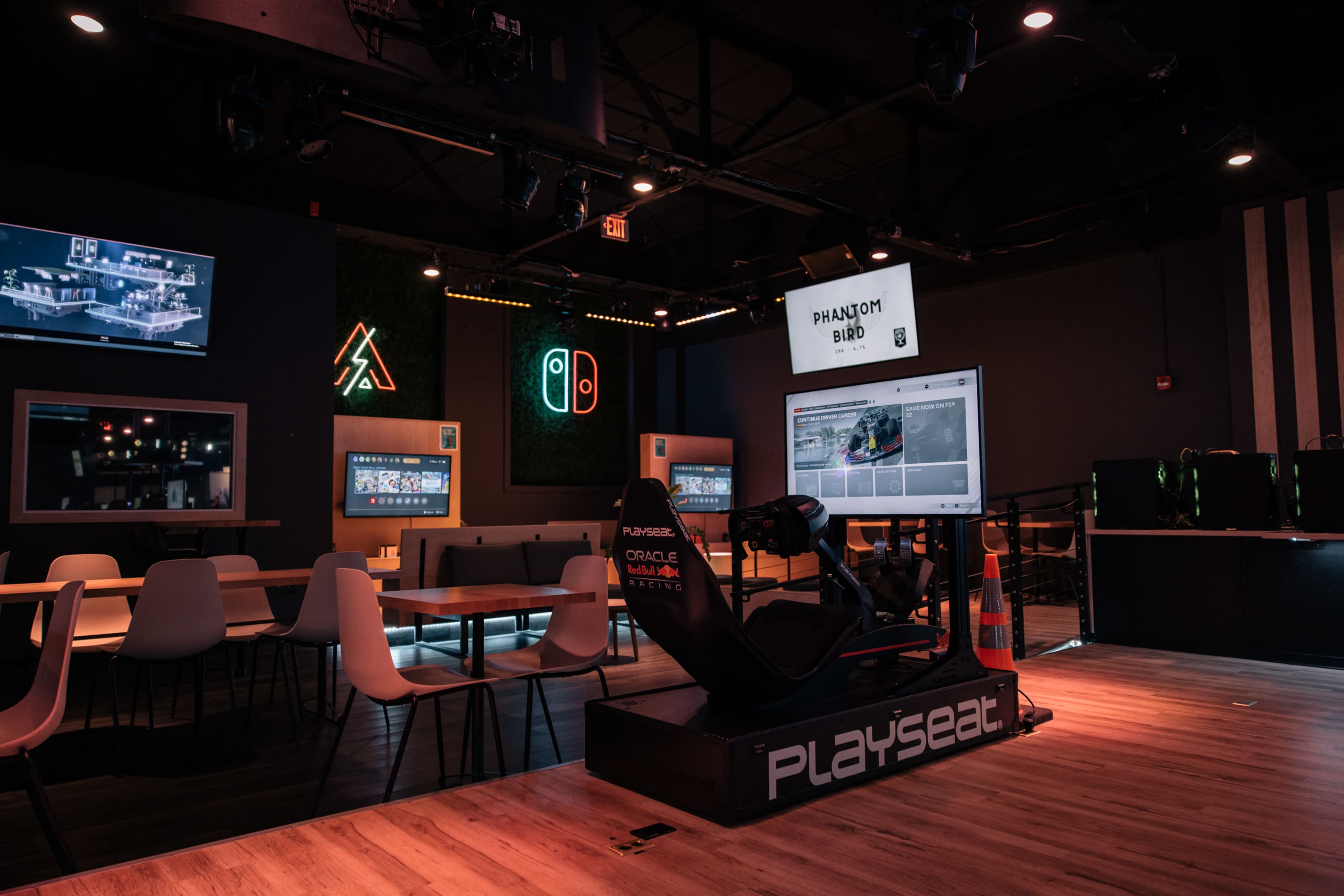 The image shows a modern gaming lounge with seating, screens displaying gaming content, and a Playseat set up for simulation racing.