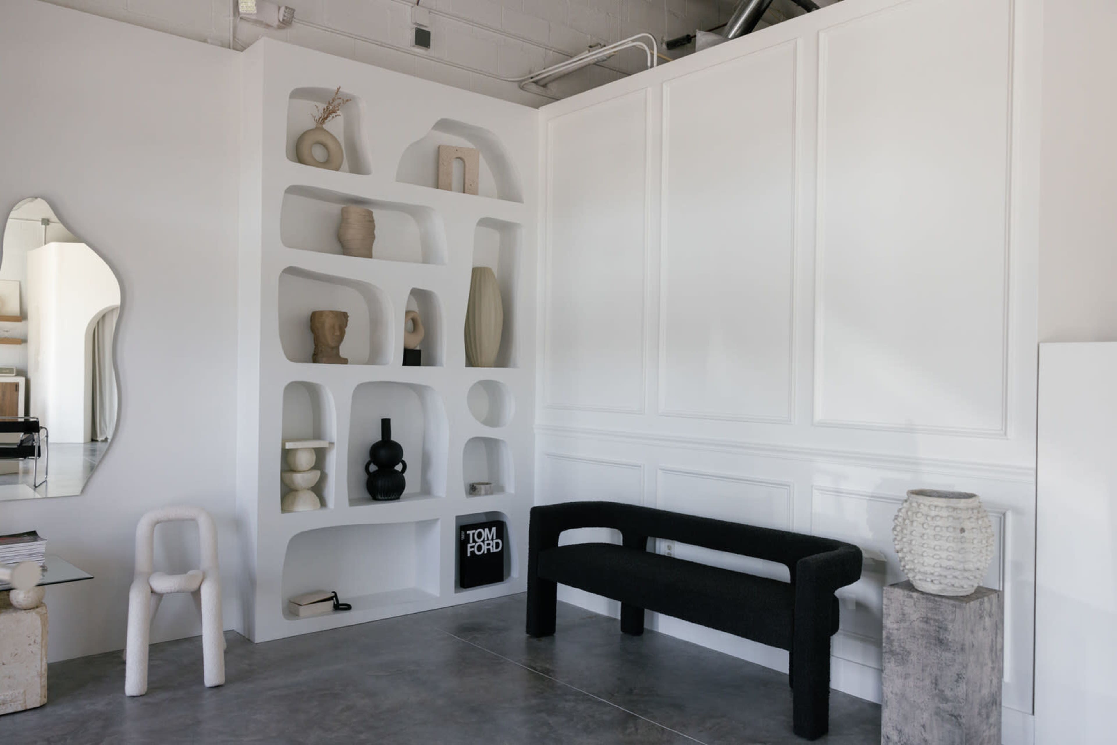 A modern interior space featuring white shelves with various sculptural objects, a black sofa, a round mirror, and a concrete floor.