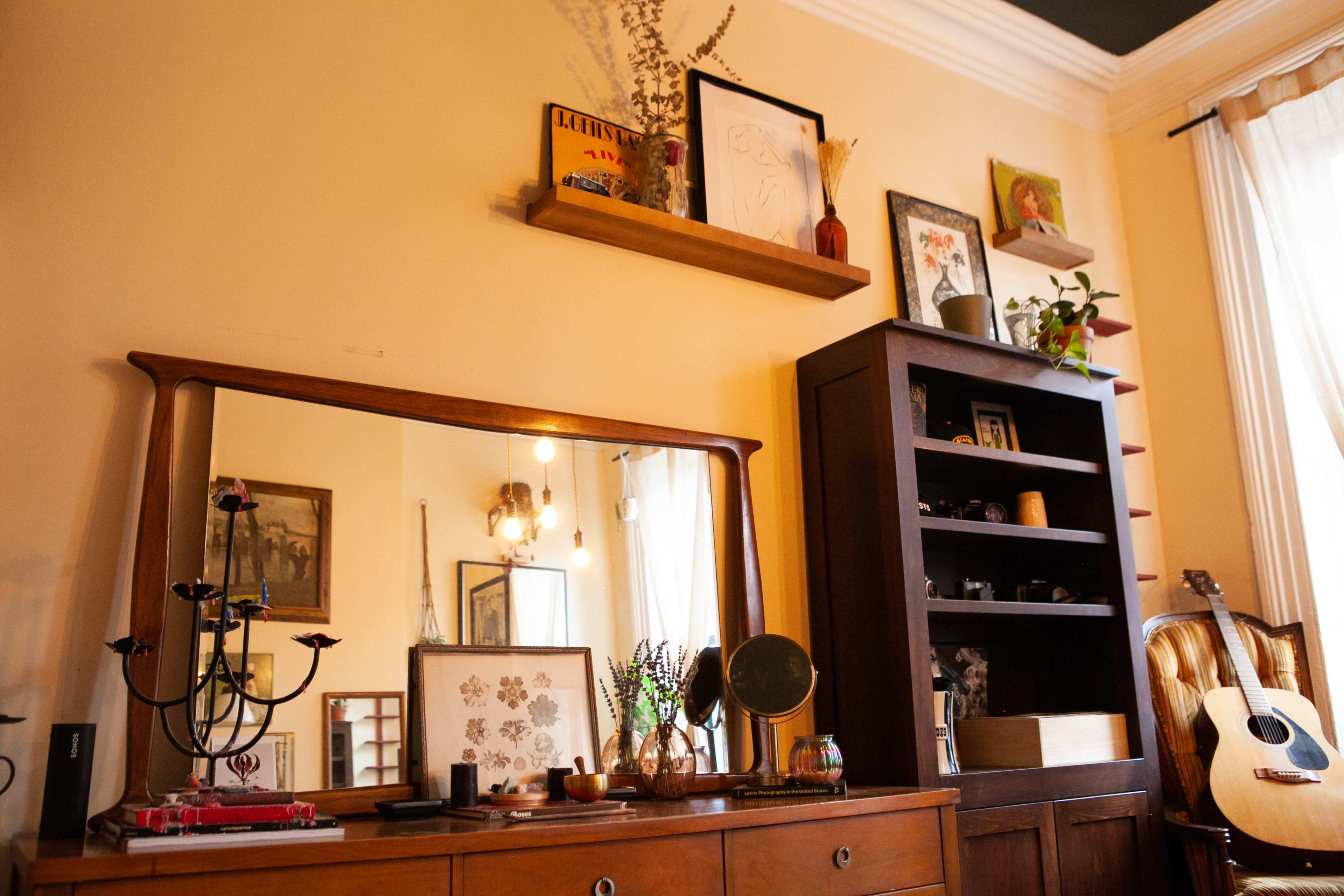 The image shows a cozy living space featuring a wooden dresser with a mirror, shelves with decorative items, and a black bookshelf next to an acoustic guitar.