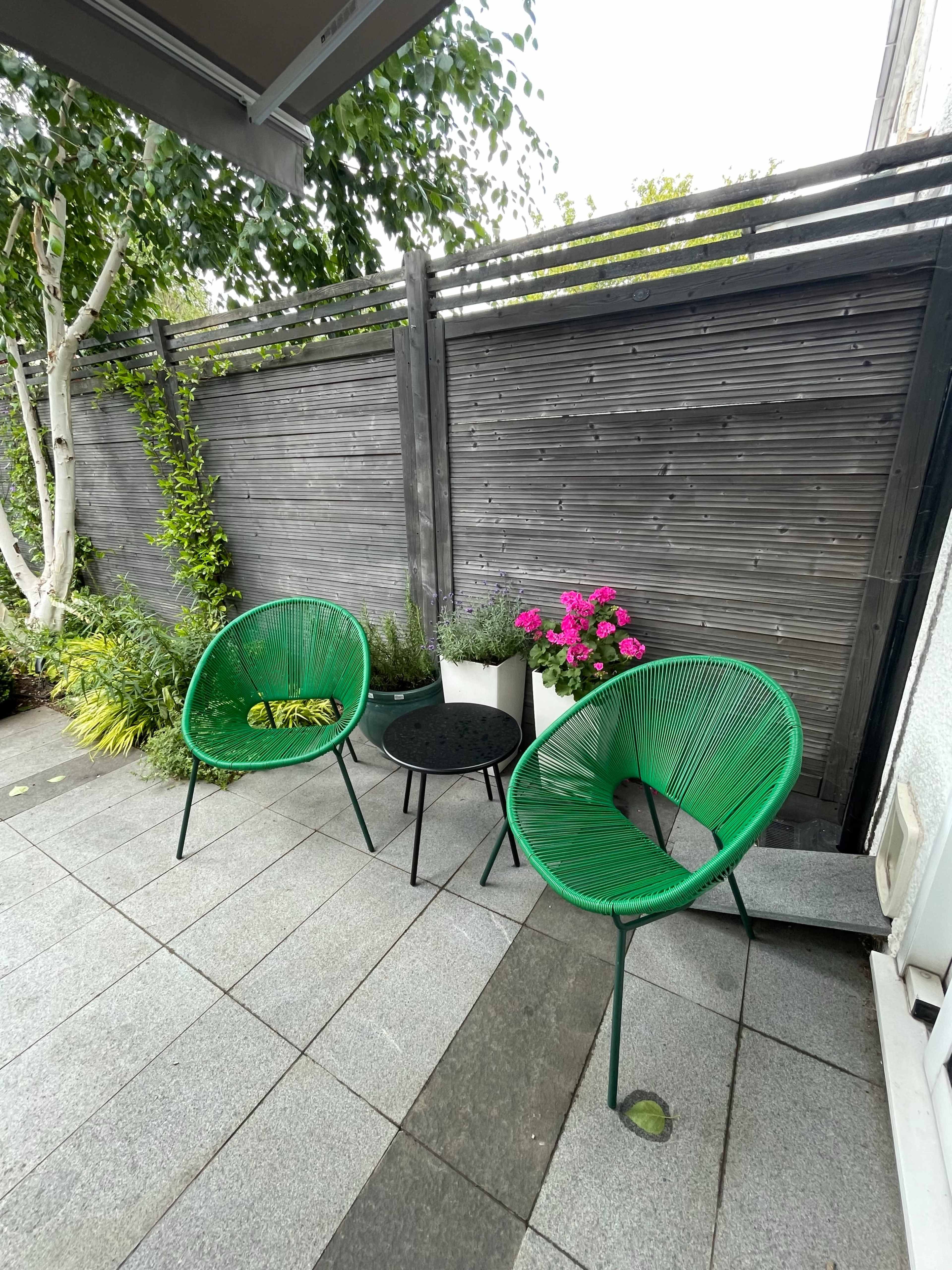 A small outdoor seating area with two green chairs, a black round table, and a potted plant with pink flowers beside a wooden fence.
