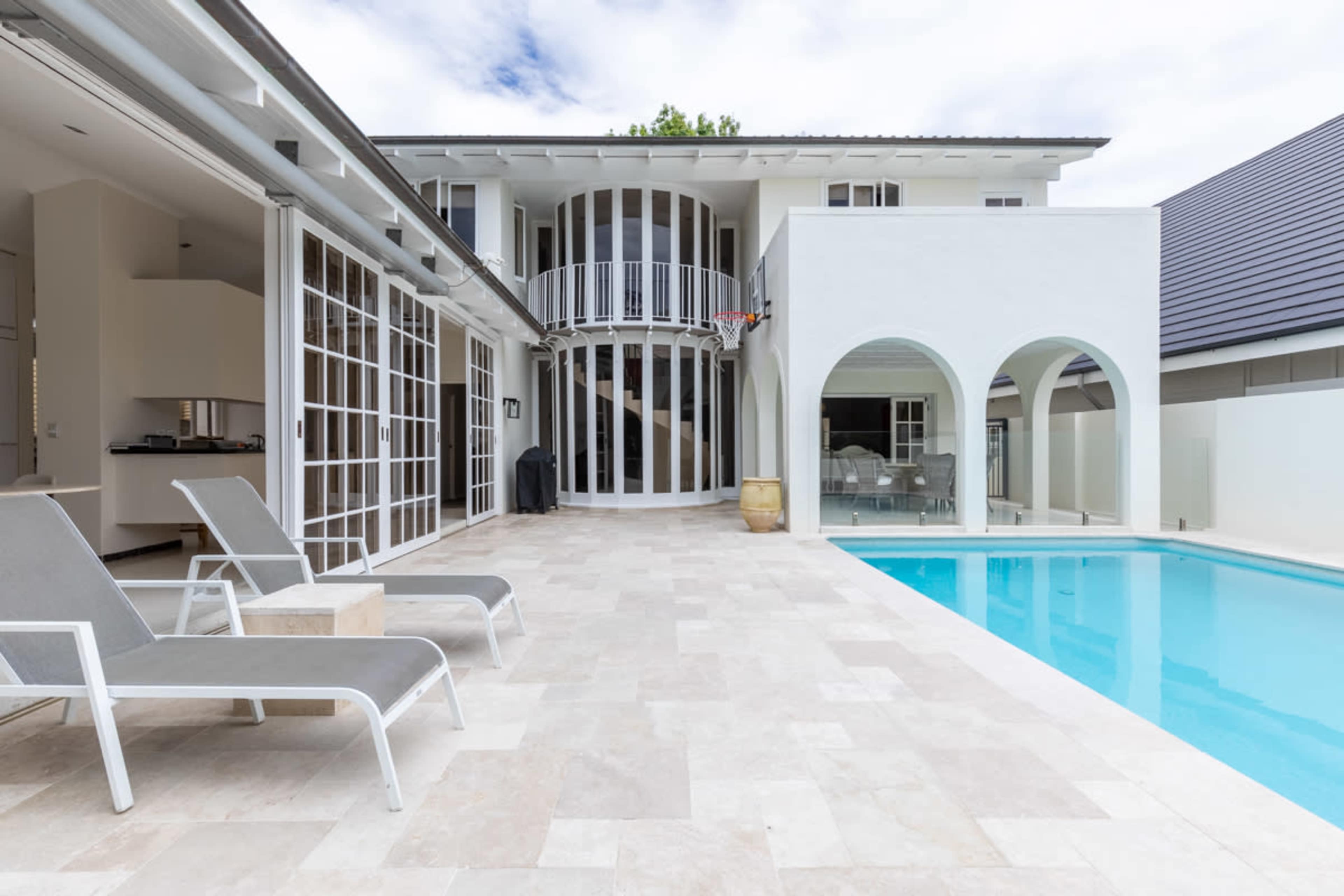 The image shows a modern backyard with a swimming pool, lounge chairs, and a multi-level house featuring large glass doors and arches.