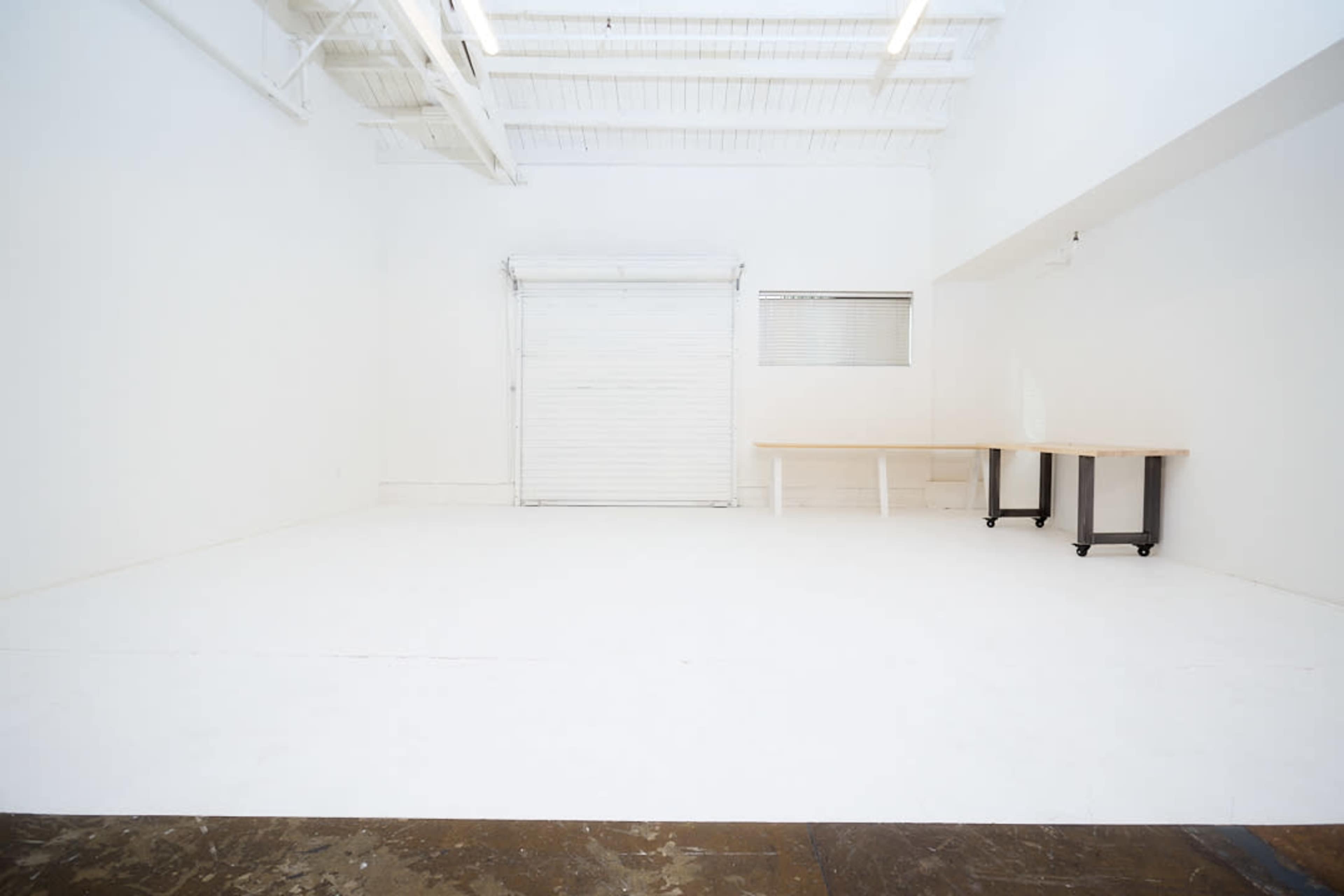 The image shows a spacious, empty room with white walls, a roll-up door, a window, and a simple wooden table on wheels.