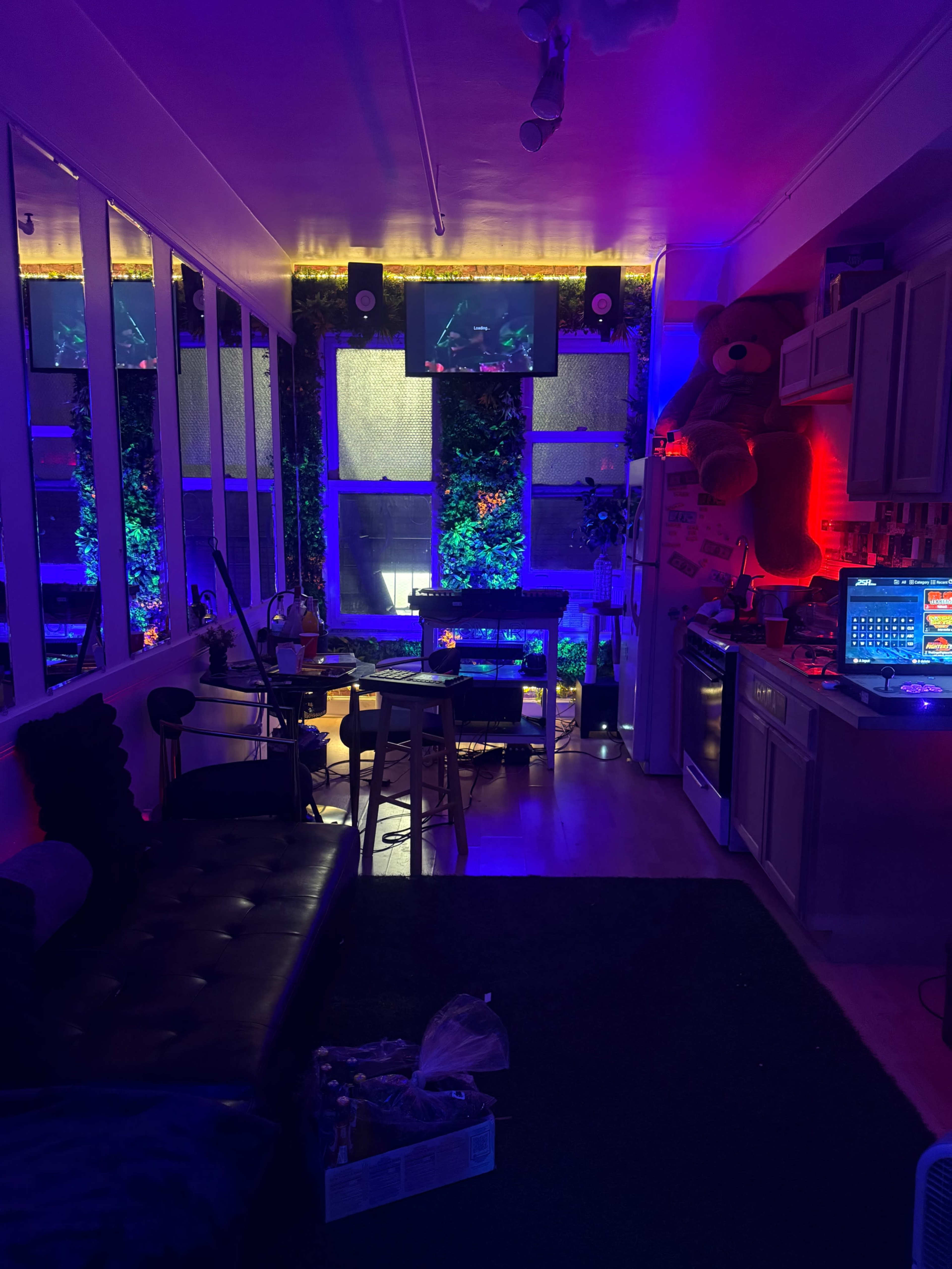 The image shows a dimly lit room with colorful LED lights, a kitchen area, a small table with chairs, and a large teddy bear in one corner.