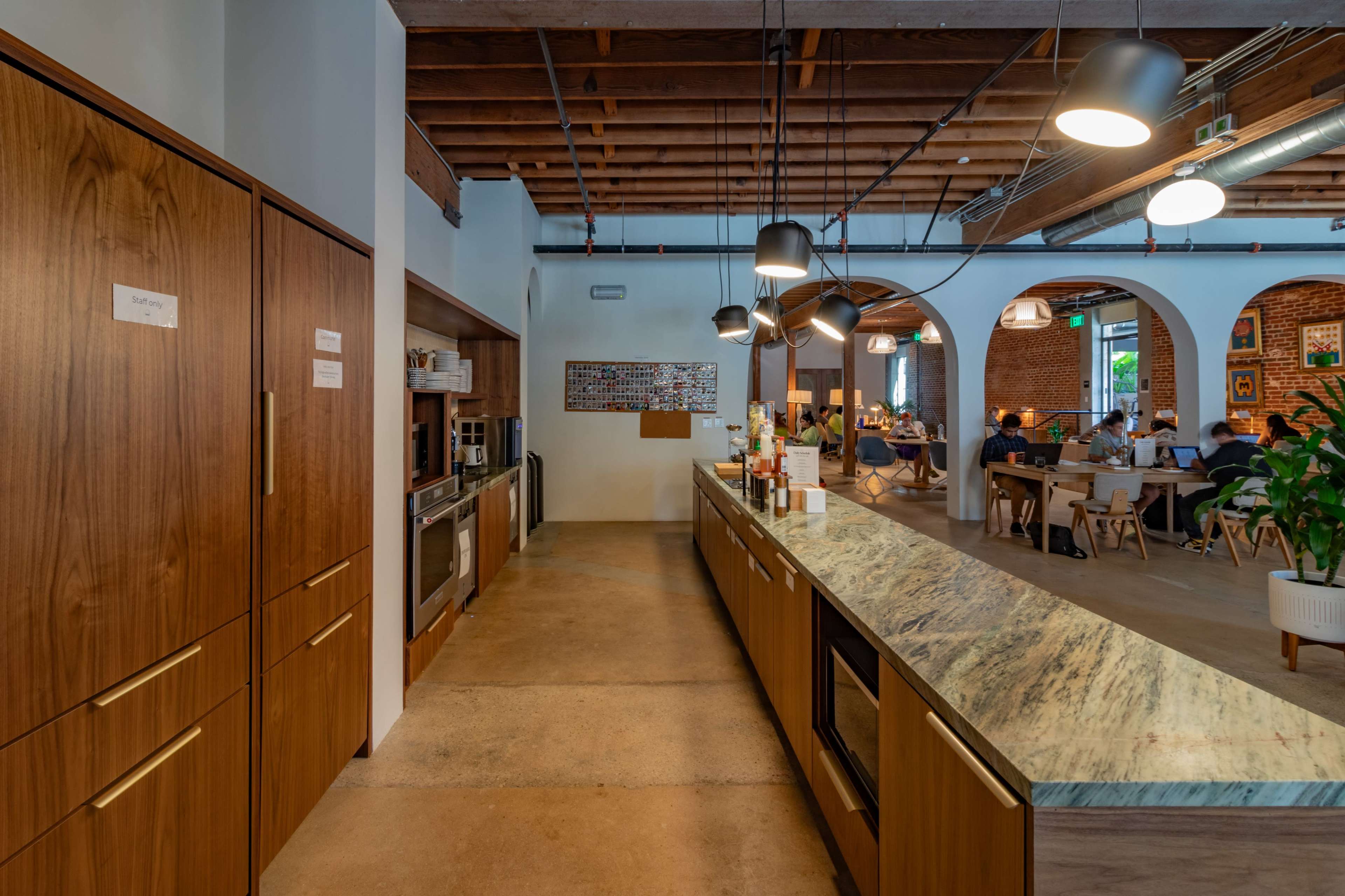 A modern kitchen area with wooden cabinetry, a marble counter, and an open layout leading to a dining space filled with people.