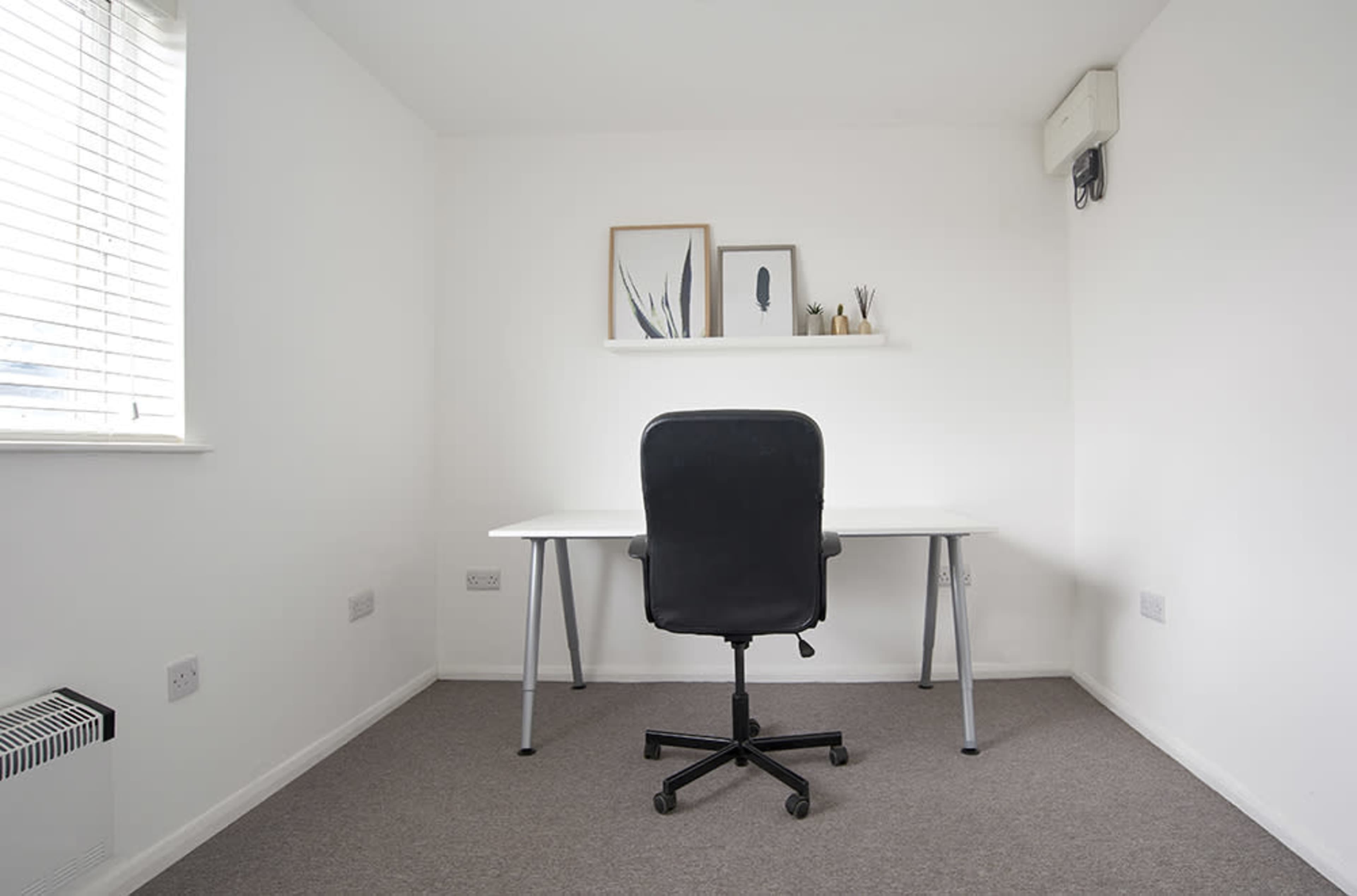 A minimalistic room with a desk and a chair, featuring a window with blinds and a small shelf on the wall.