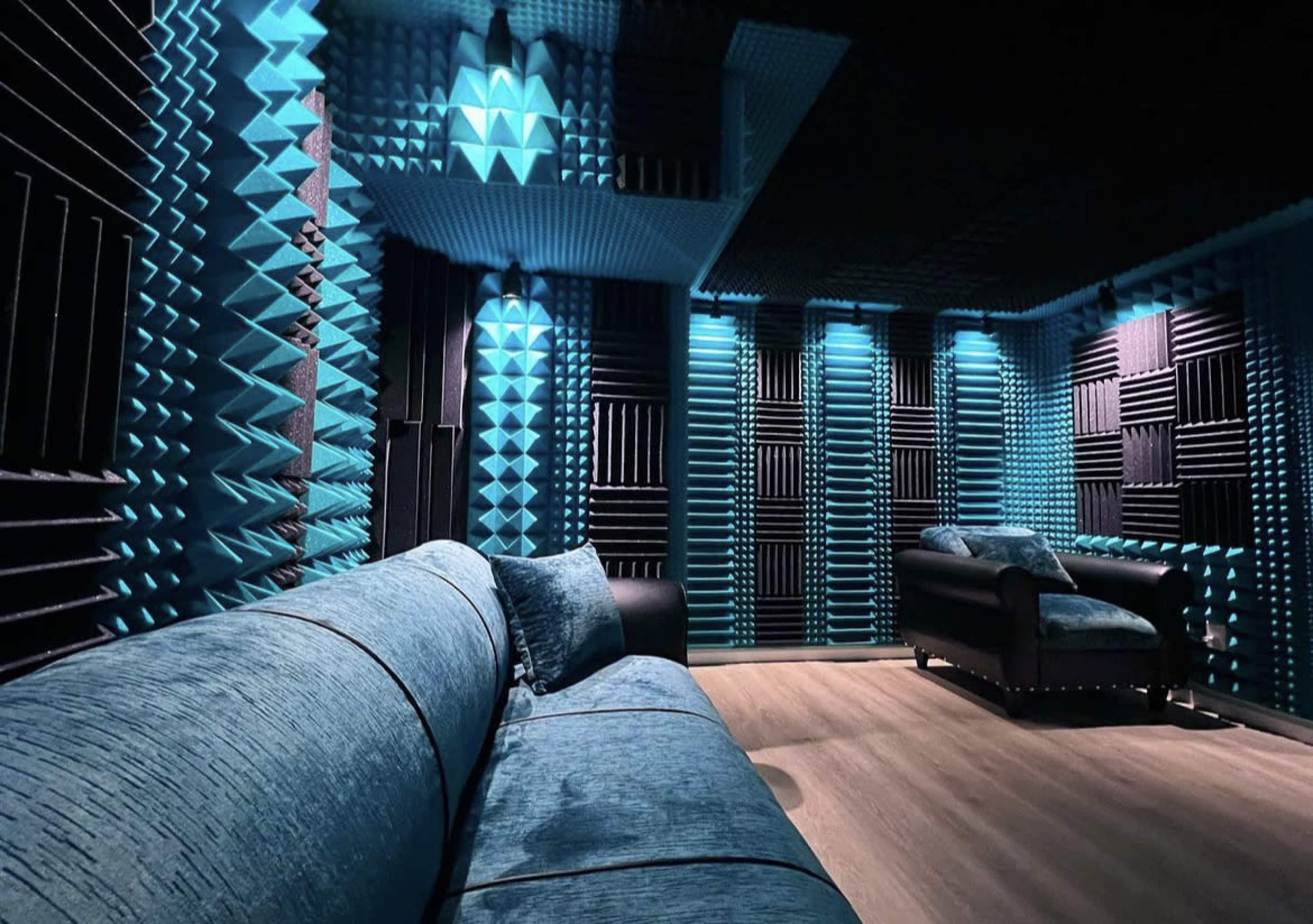 A soundproof room features blue and black acoustic panels on the walls, with a blue sofa and a black armchair arranged on a wooden floor.