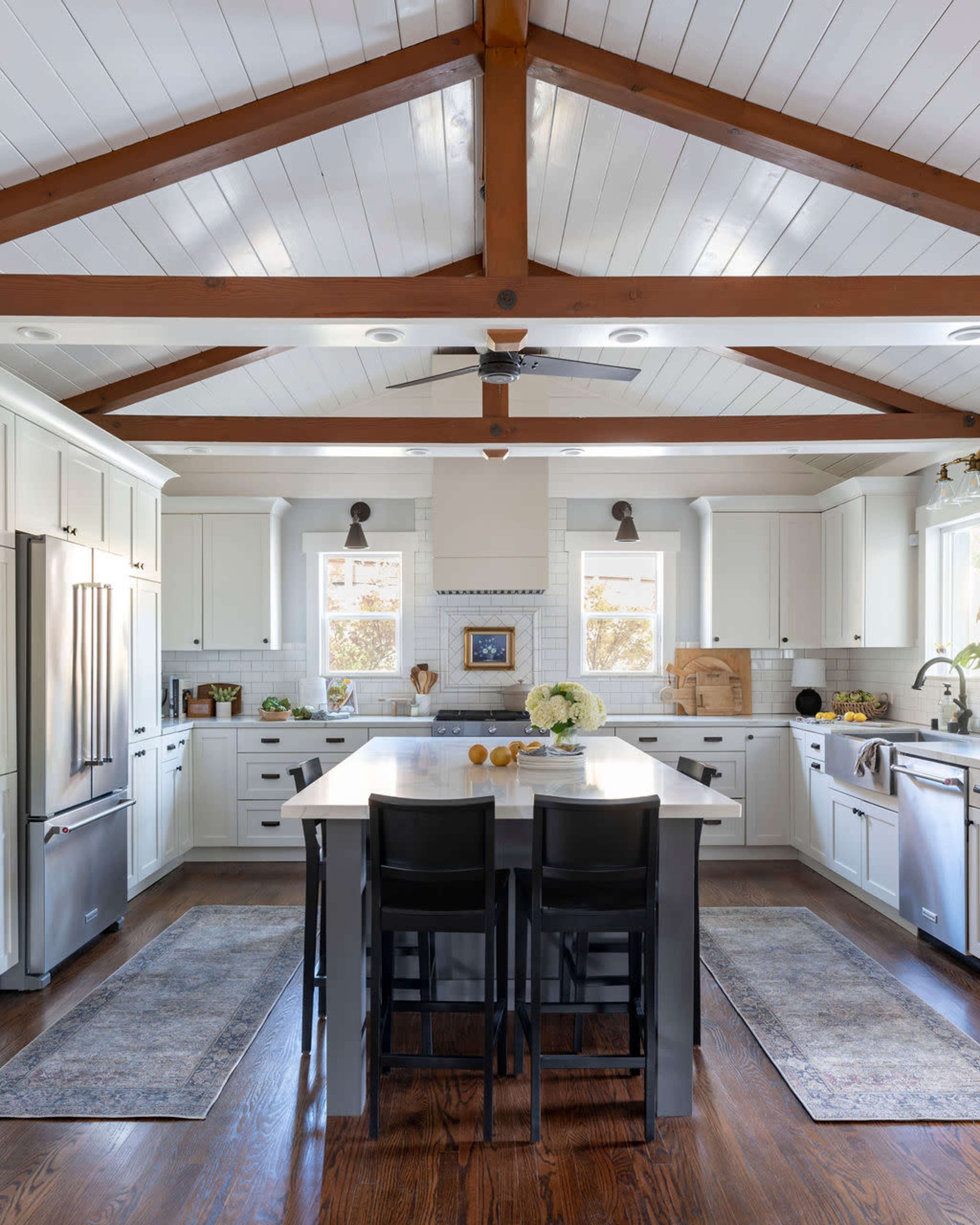 A modern kitchen features white cabinetry, a large island with dark seating, and wooden ceiling beams.