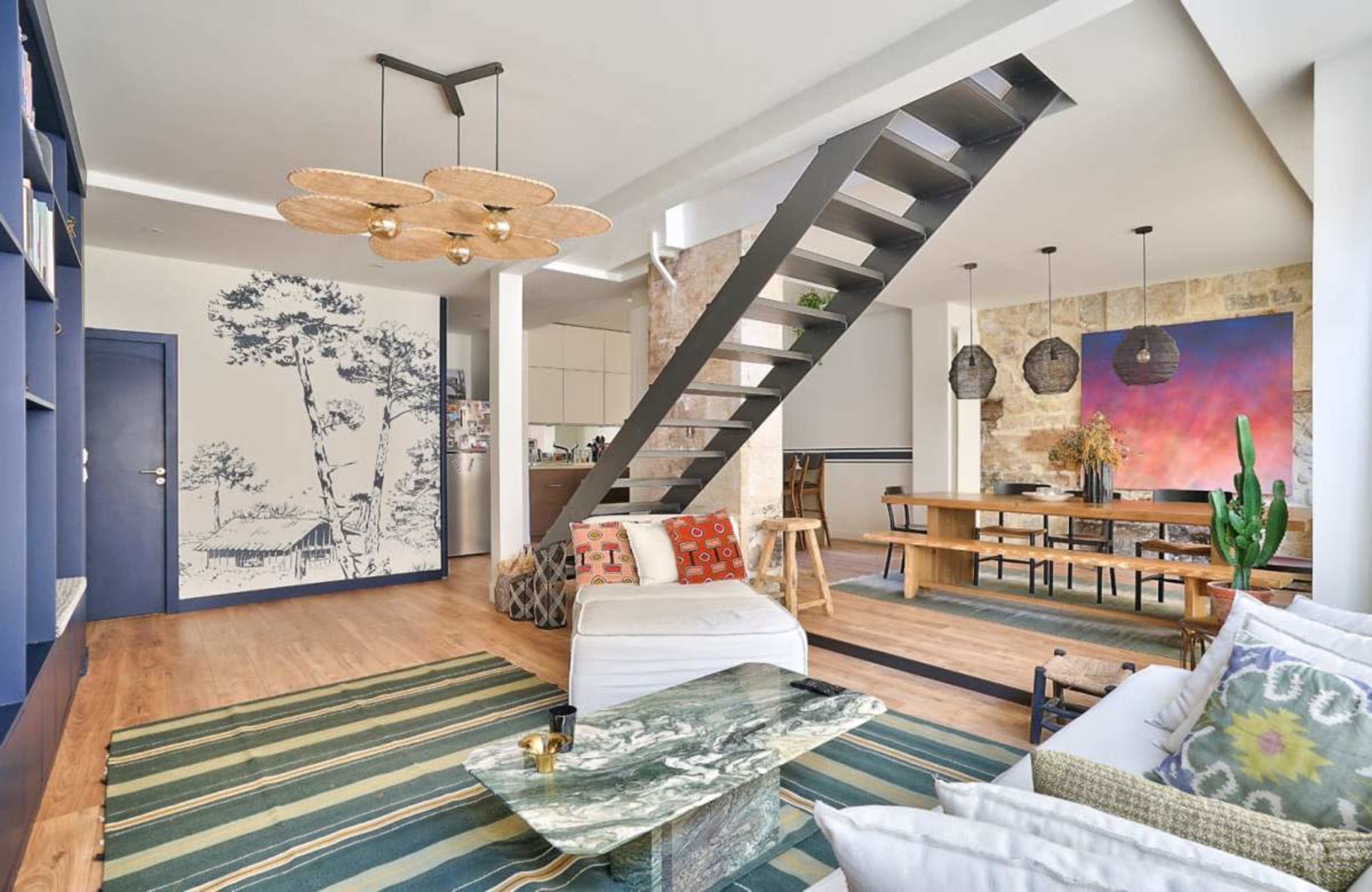 The image shows a modern living space featuring a staircase, a seating area with decorative cushions, a large dining table, and various artistic elements on the walls.