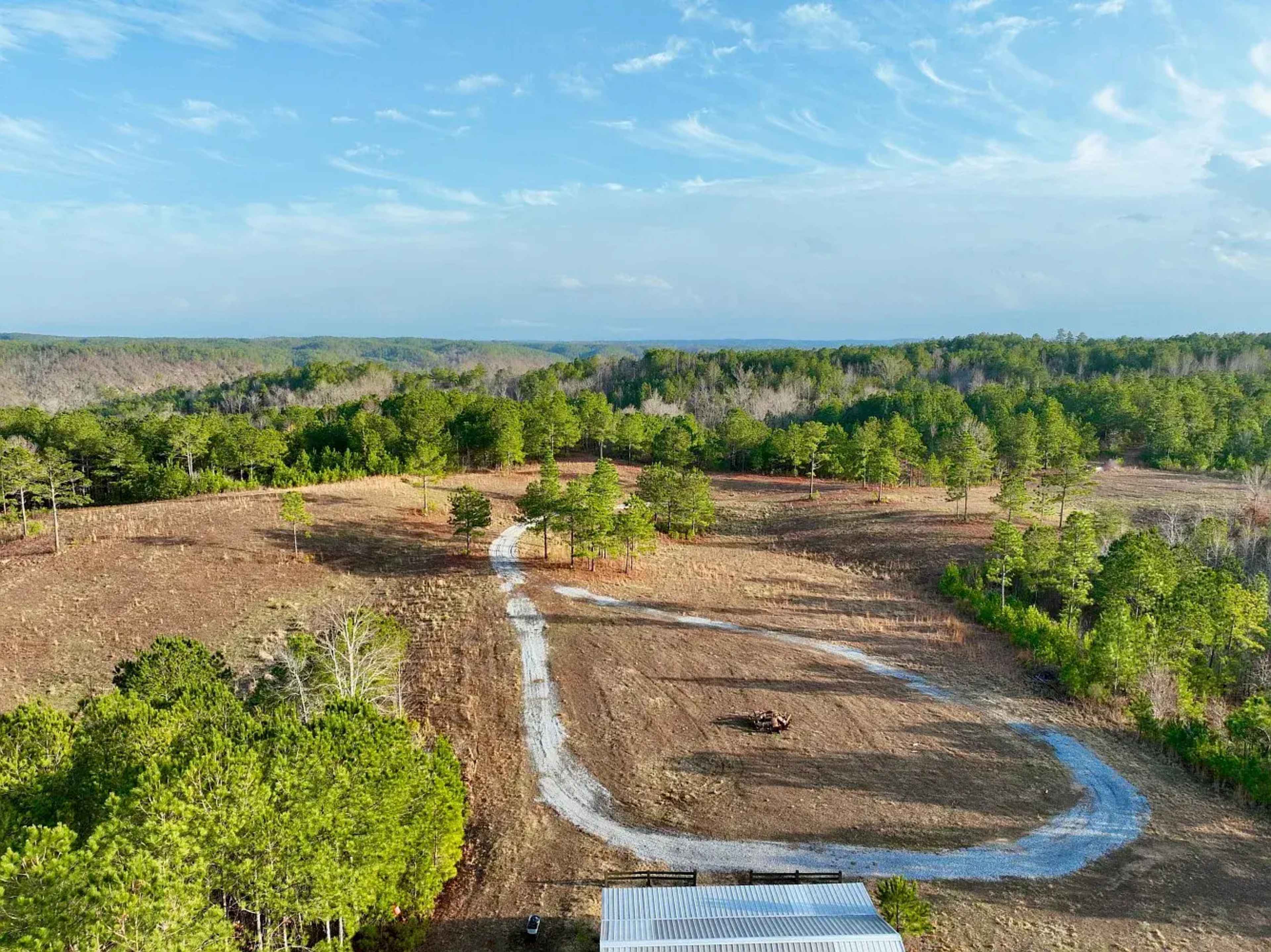 The image shows an aerial view of a cleared landscape with a winding dirt path leading through scattered trees under a partly cloudy sky.