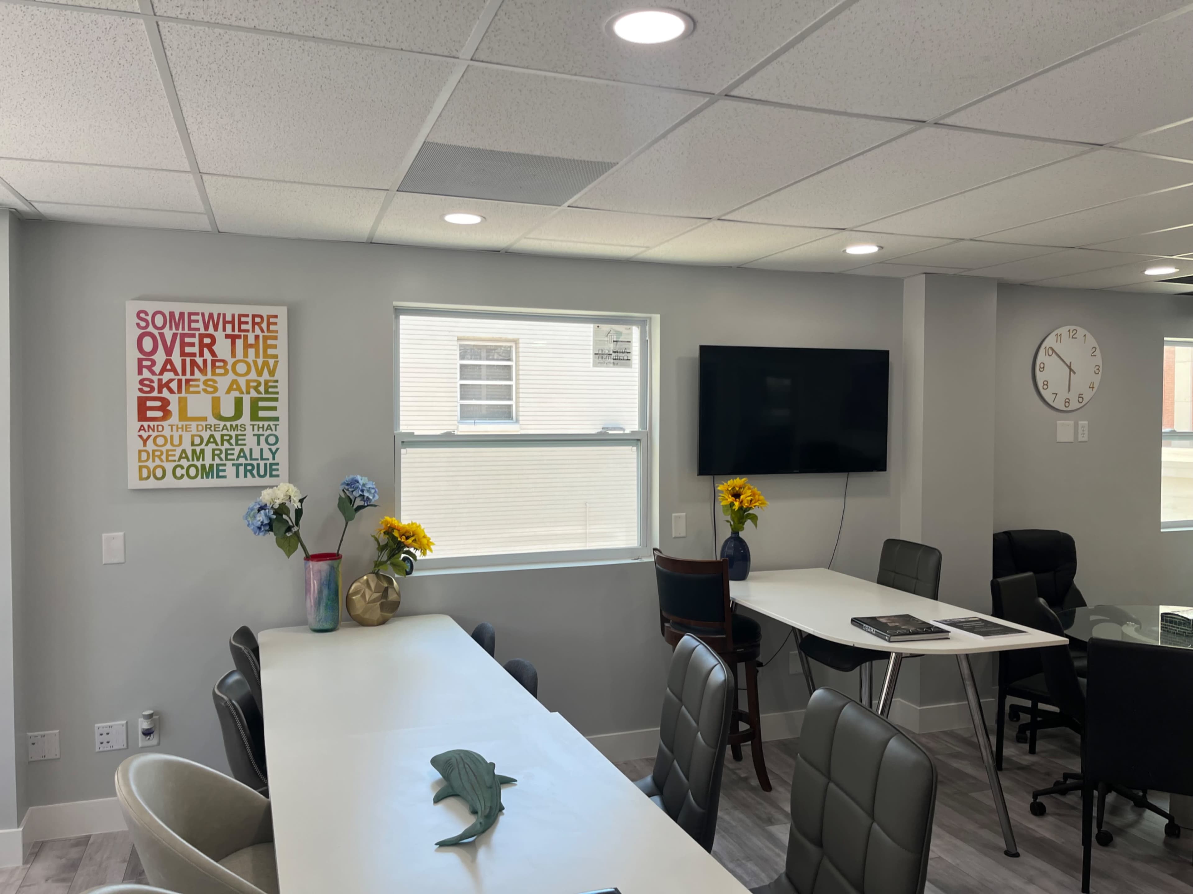 A bright and modern office space features several tables, a television on the wall, and a colorful motivational poster.
