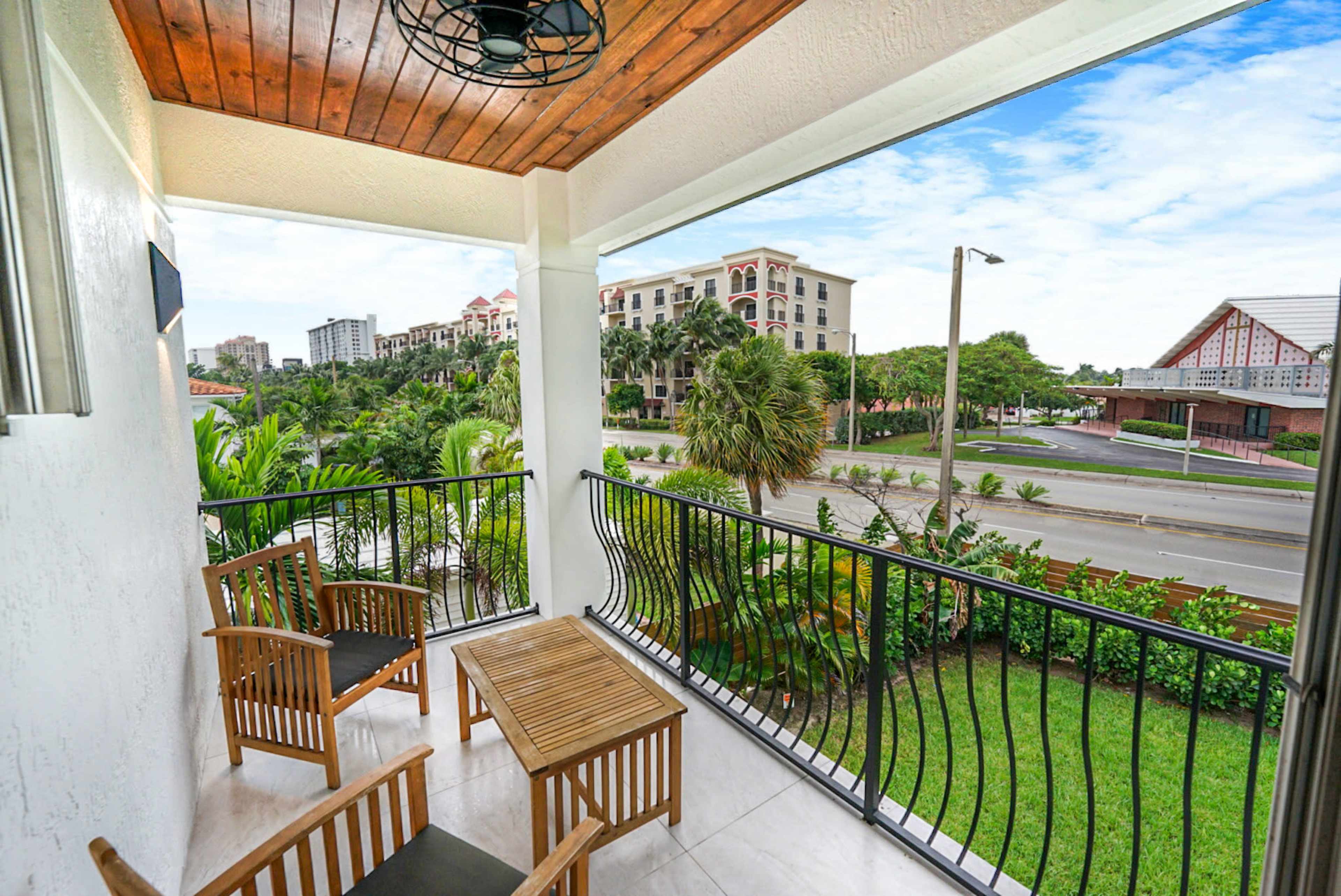 The image shows a balcony with two wooden chairs and a small table overlooking a landscaped area and a road lined with buildings.