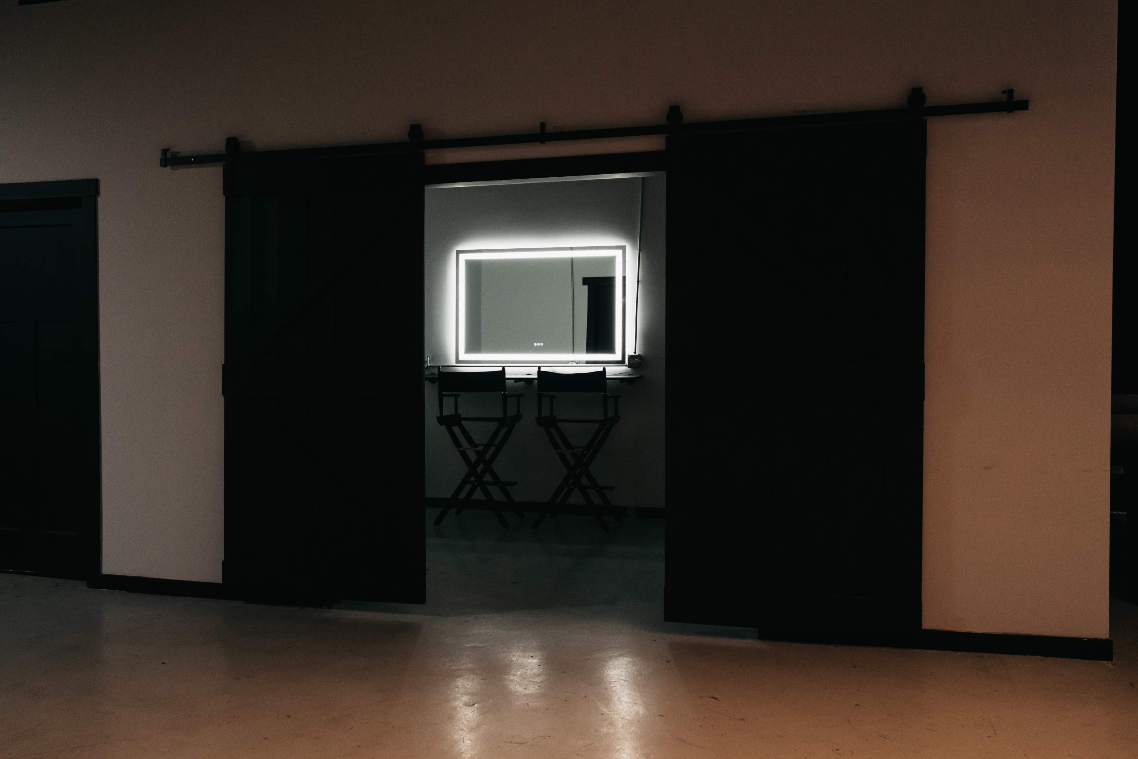 A doorway with sliding barn doors reveals a room containing a mirror with backlighting and two director-style chairs.