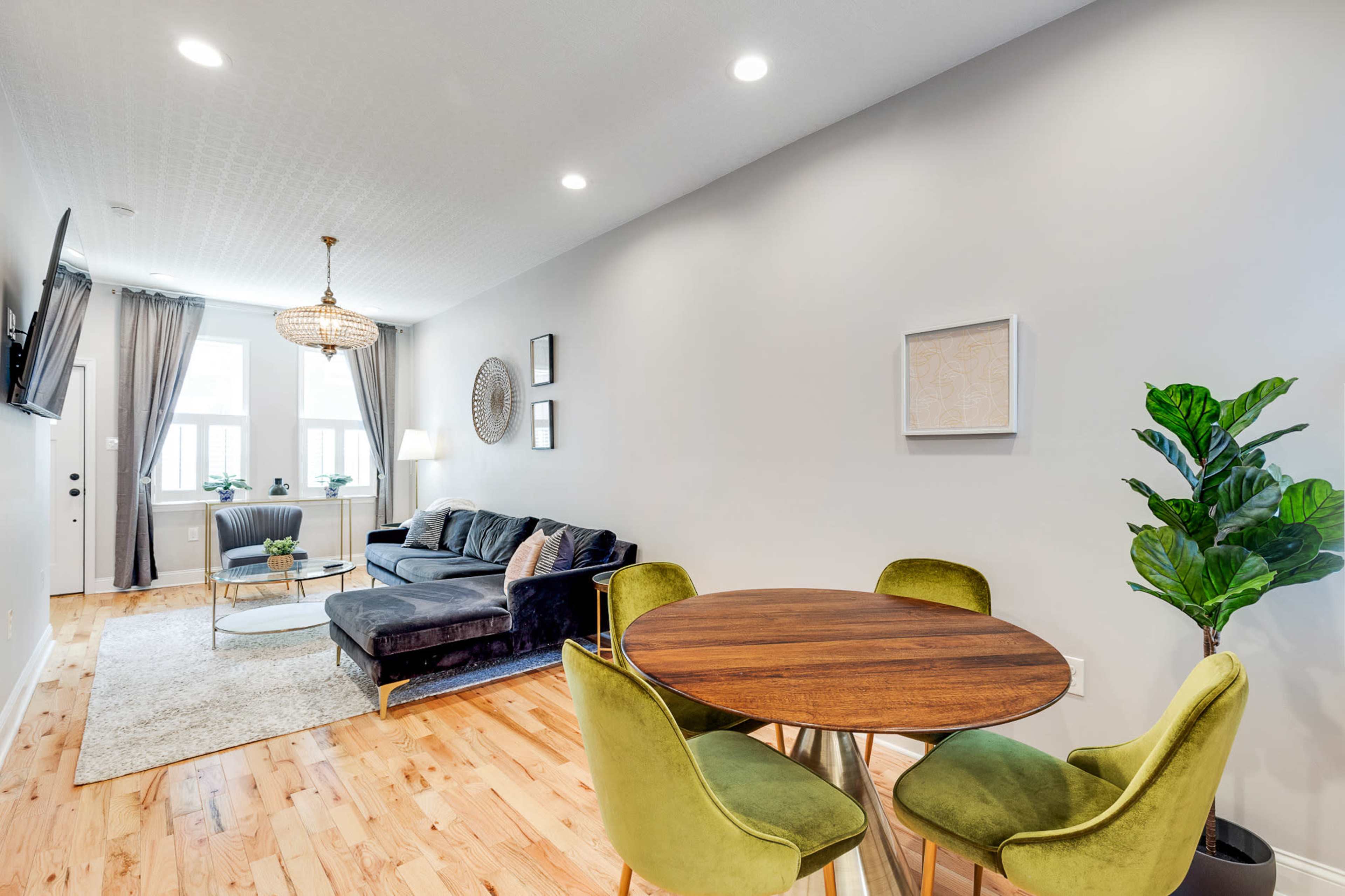 The image shows a modern living space featuring a dark sofa, a round wooden dining table with four green chairs, and a potted plant against a light gray wall.