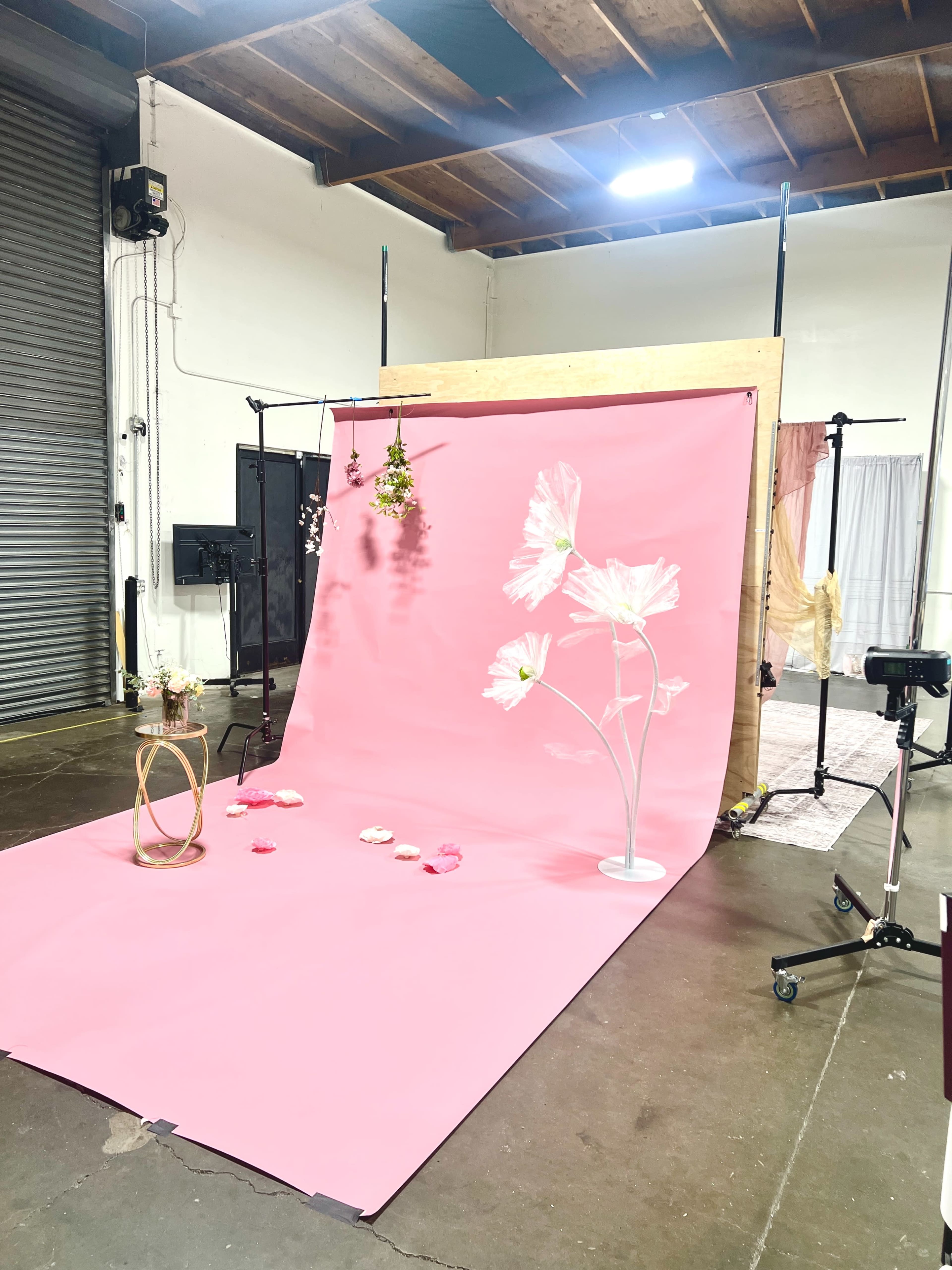 The image shows a photography setup featuring a pink backdrop with large artificial flowers, a small wooden table, and floral decorations scattered on the floor.