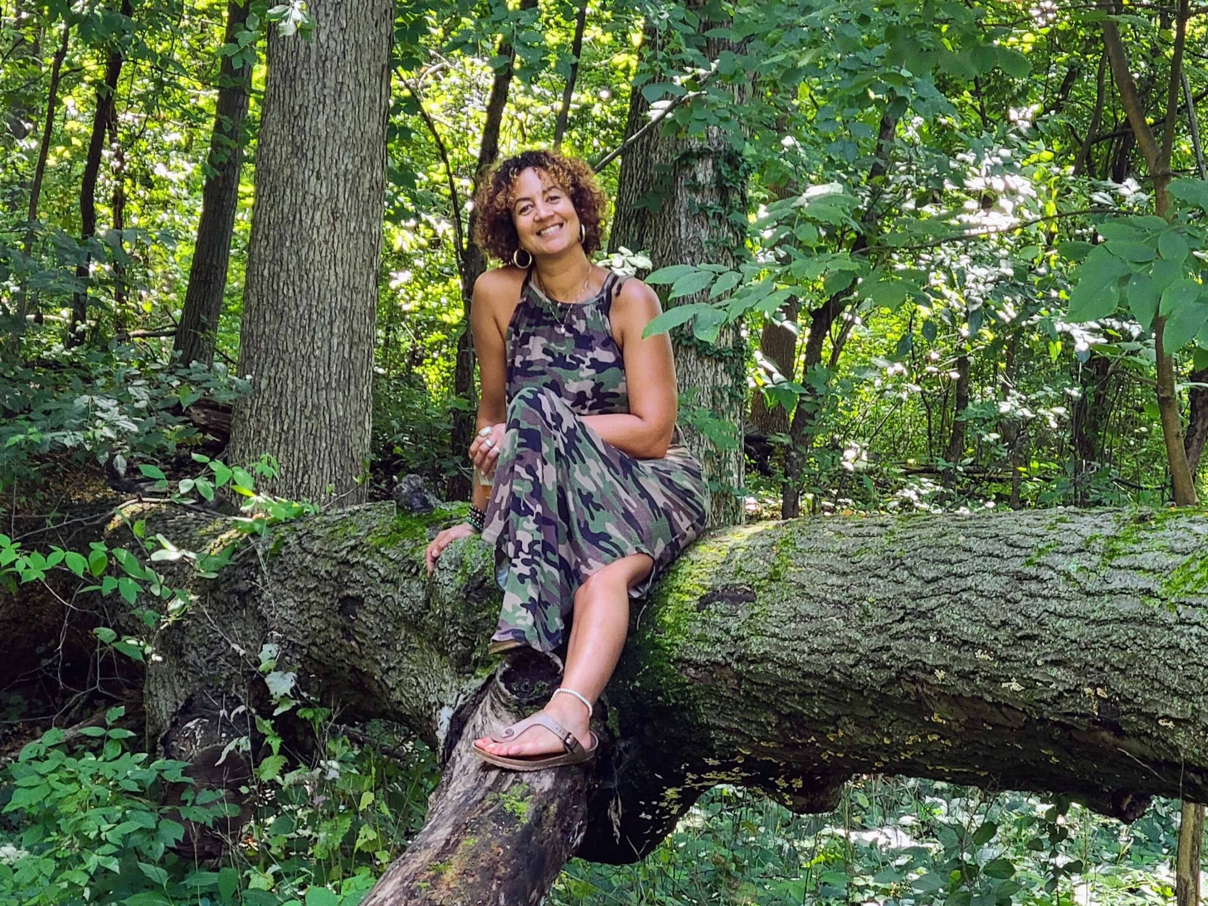 A woman wearing a camouflage dress sits on a fallen tree in a wooded area surrounded by greenery.