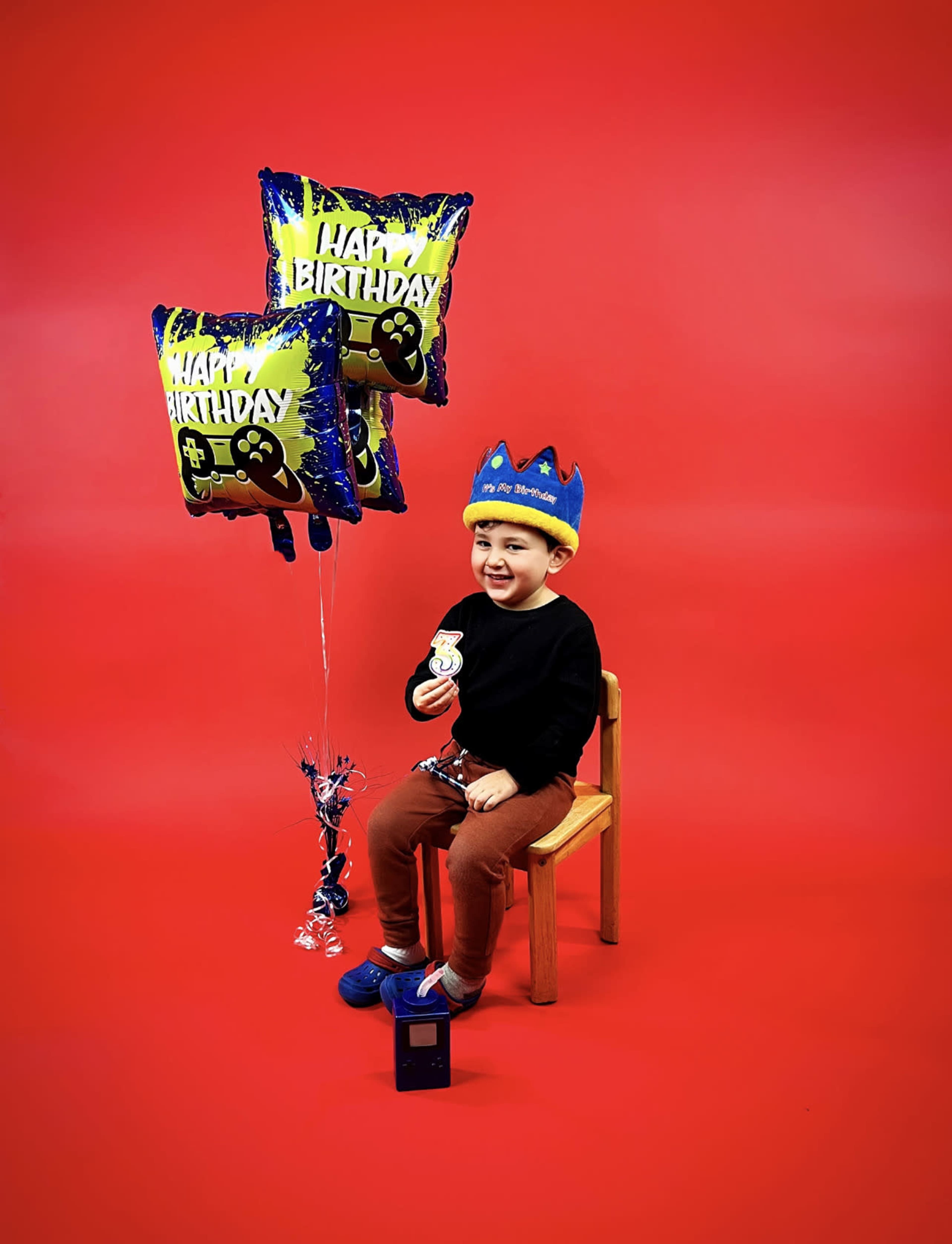A young child sits on a wooden chair holding birthday balloons and a cupcake against a red background.