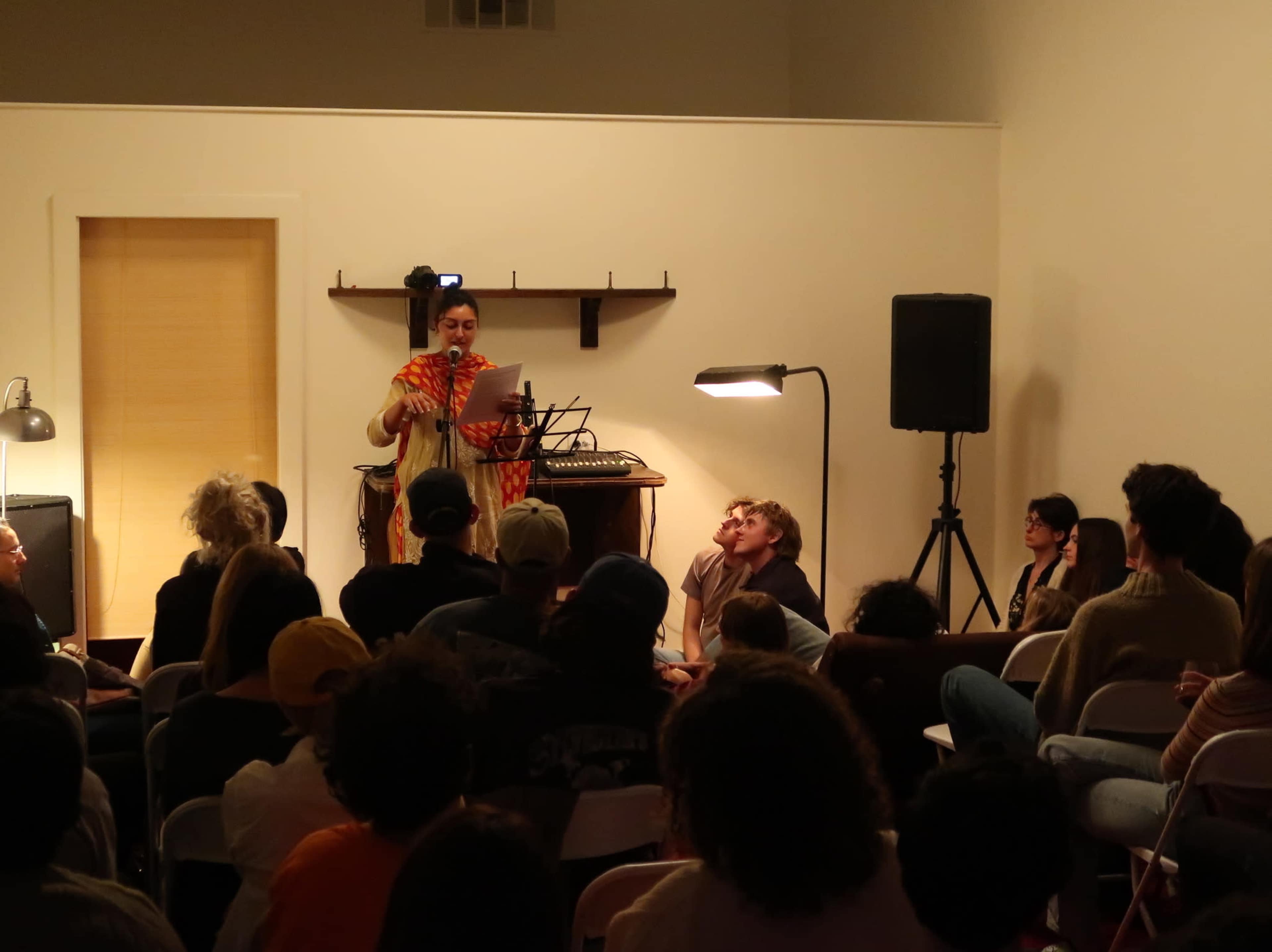 A performer reads from a script in front of an audience seated on the floor and chairs in a warmly lit room.