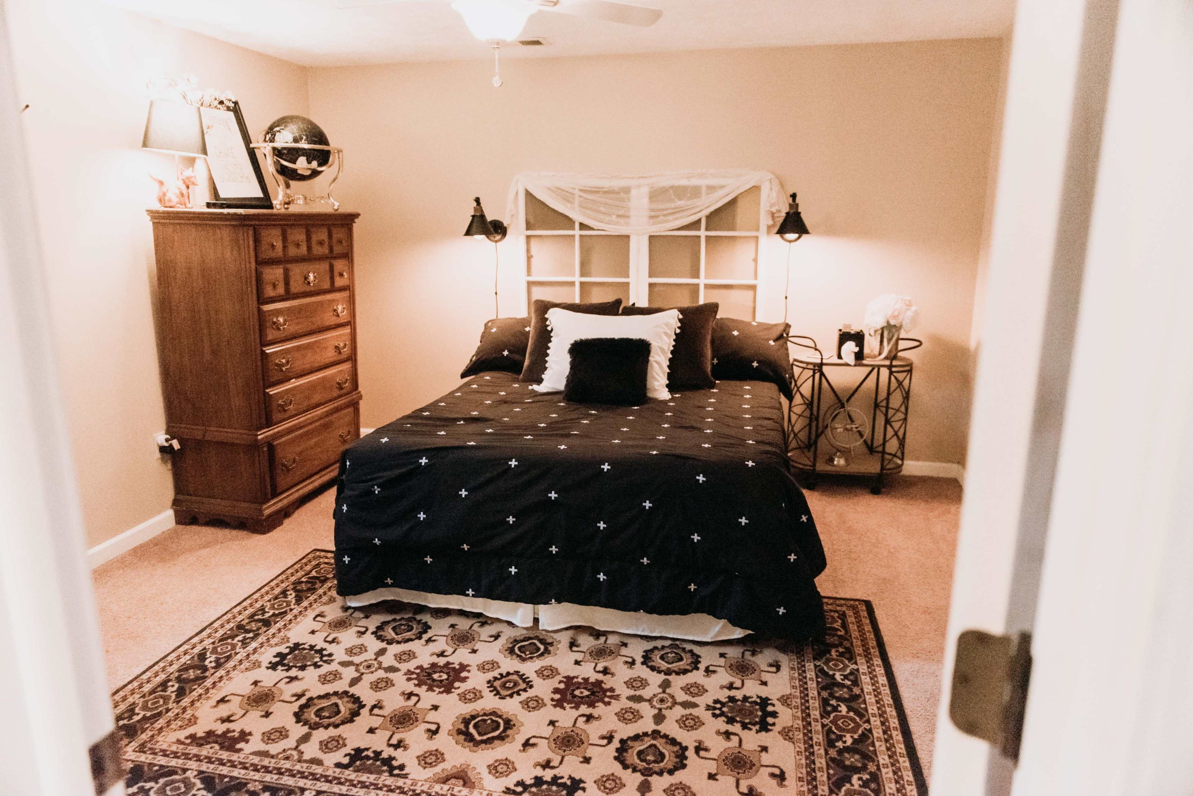 A bedroom features a bed with a dark patterned comforter, a wooden dresser, and a decorative rug on the carpeted floor.