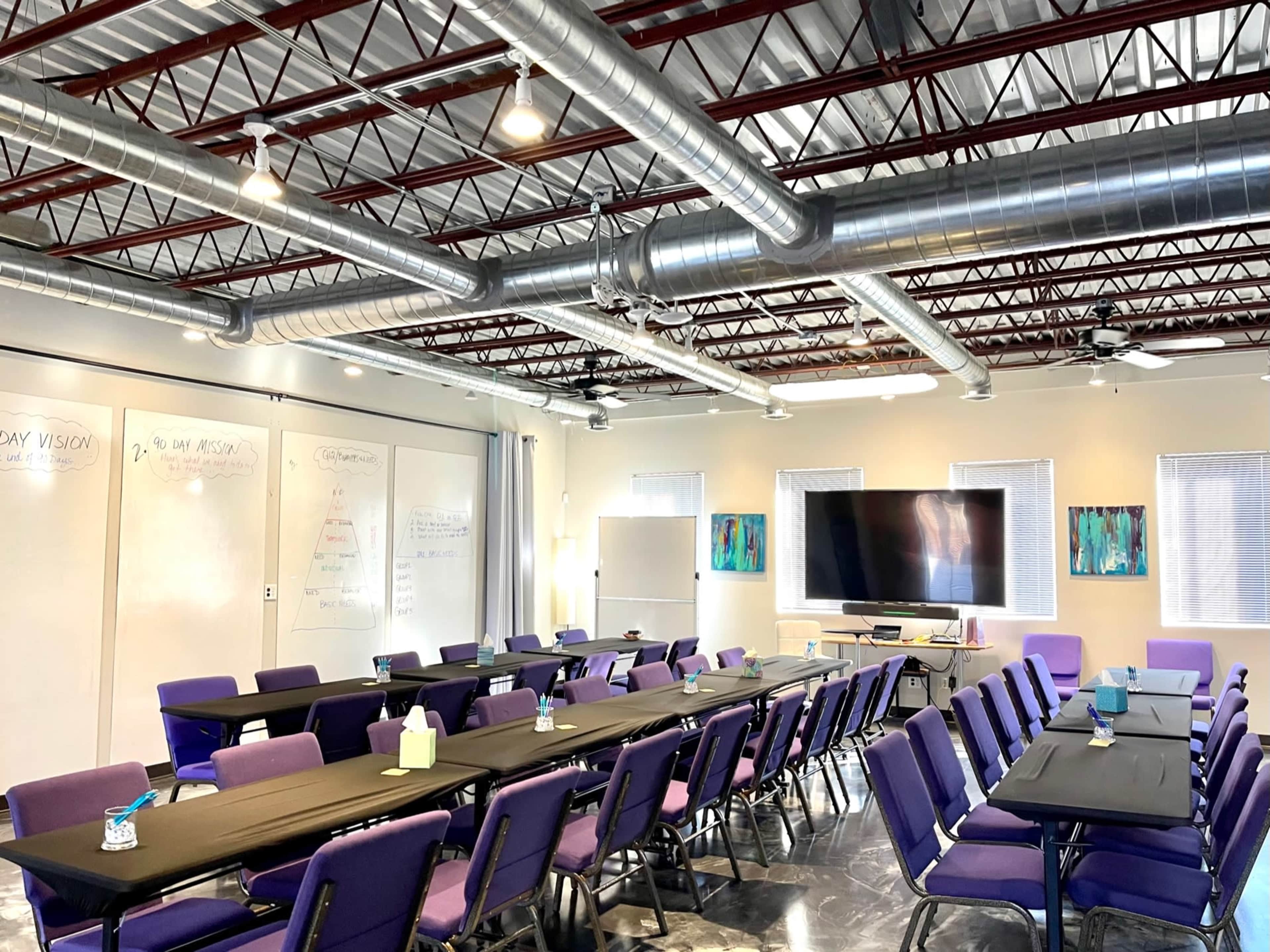 A spacious conference room is set up with multiple tables and purple chairs for a meeting, featuring whiteboards, a large screen, and visible ceiling ducts.