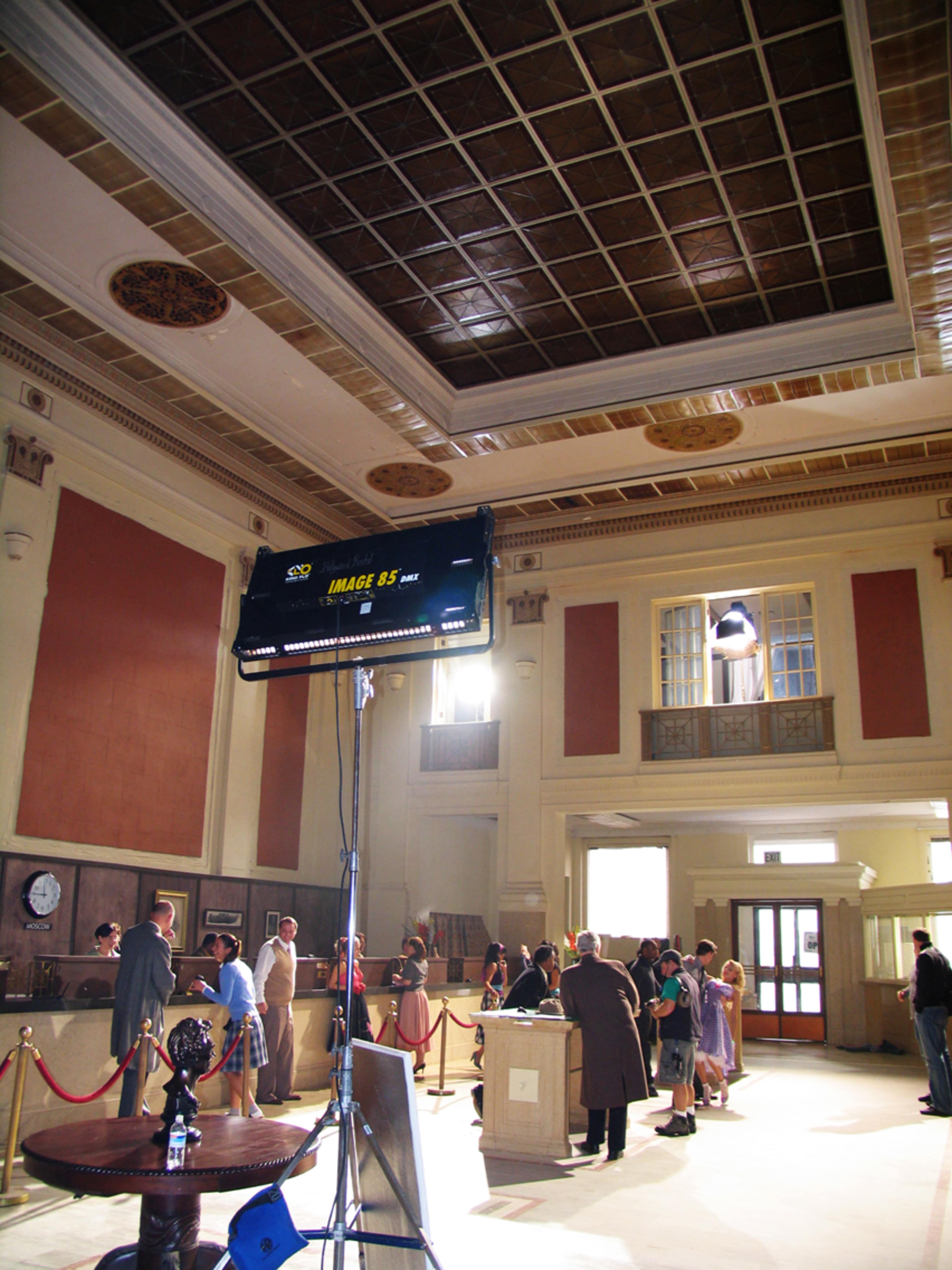 A film crew sets up a scene inside a grand, partially lit hall featuring high ceilings and decorative architecture.