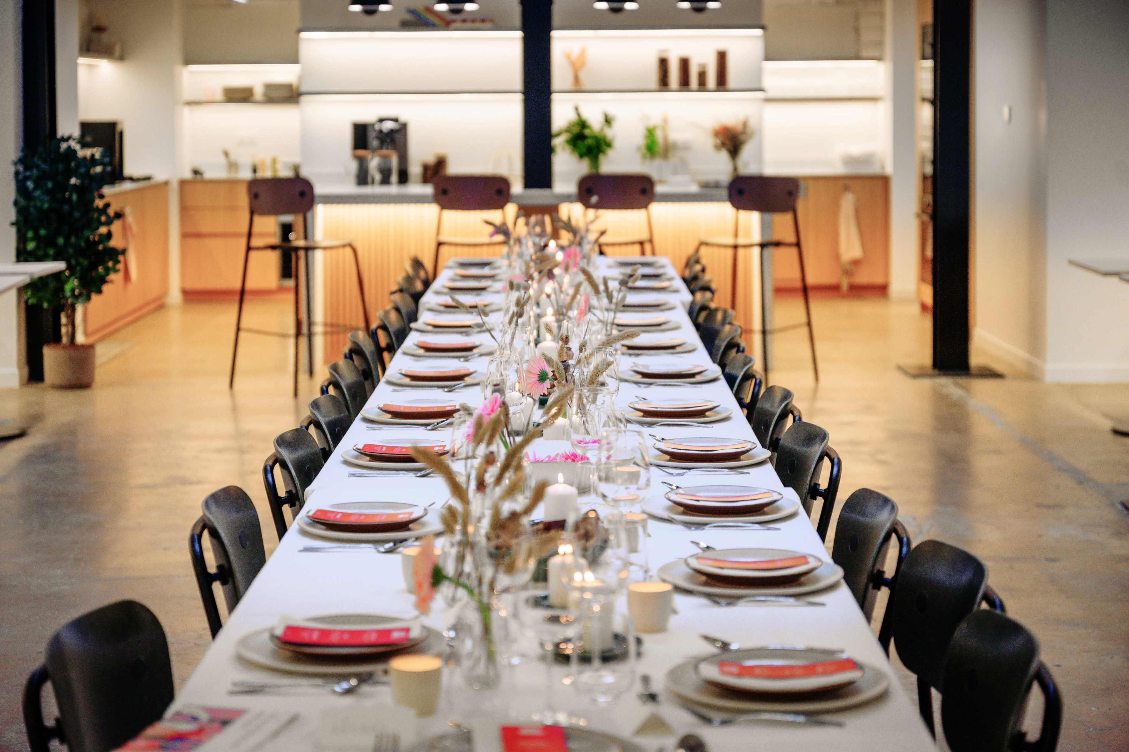 A long dining table is set with plates, glasses, and decorative centerpieces, surrounded by chairs in a bright, modern kitchen space.