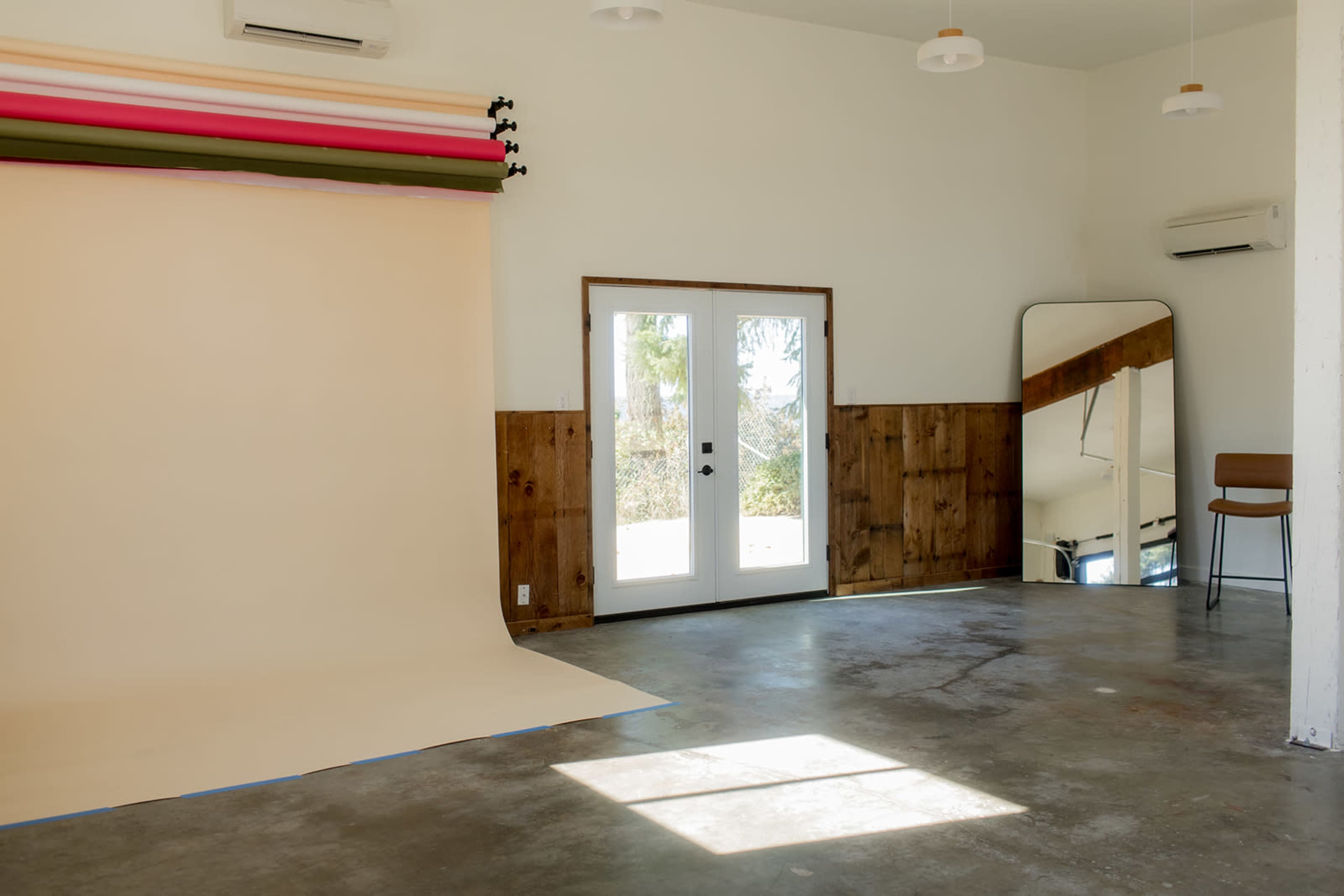 The image shows a spacious room with a beige backdrop, large windows, a wooden wall, a mirror, and a single chair.