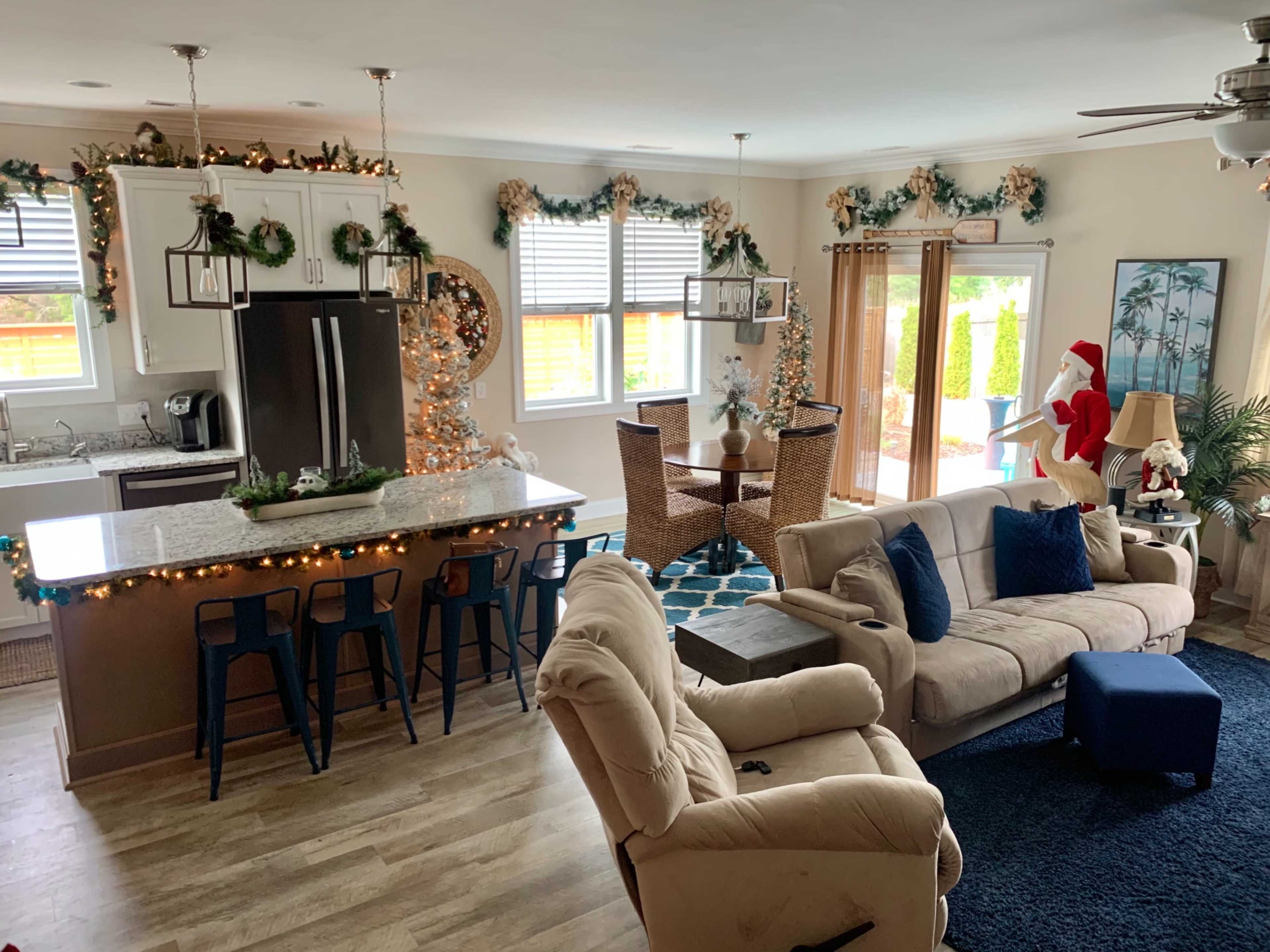 A well-decorated open-concept living space features a kitchen with bar seating, a dining area with wicker chairs, and festive holiday decorations throughout.