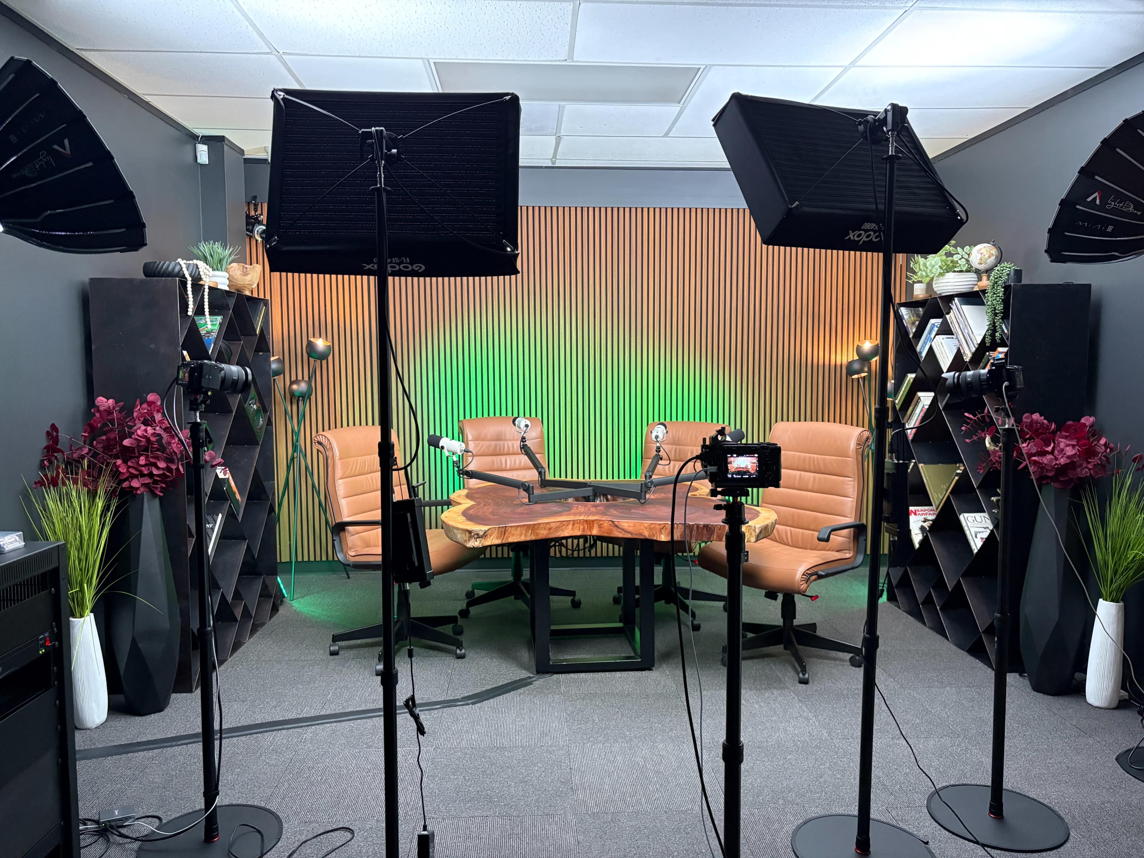 The image shows a well-lit studio setup with two brown chairs facing a wooden table, surrounded by decorative plants and shelving units.