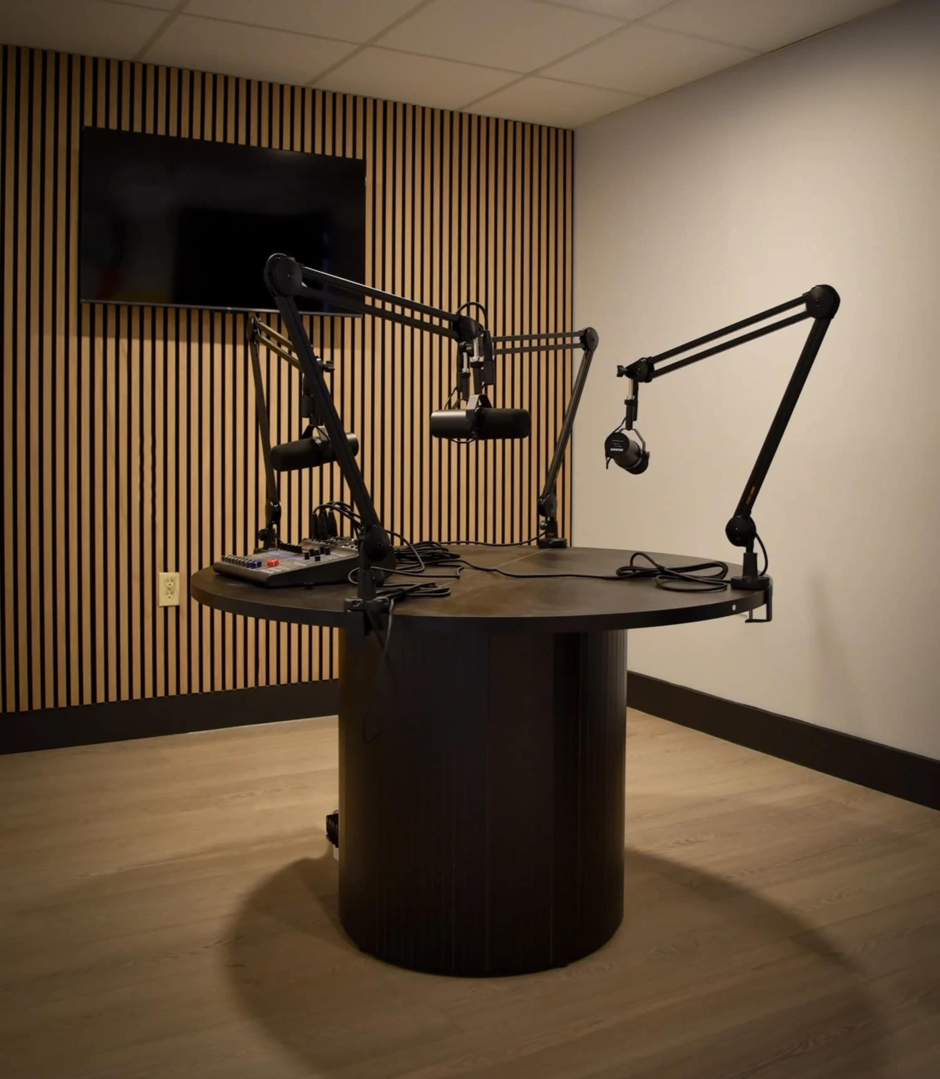The image shows a radio studio featuring a round table with adjustable microphone arms, headphones, and a monitor on the wall.
