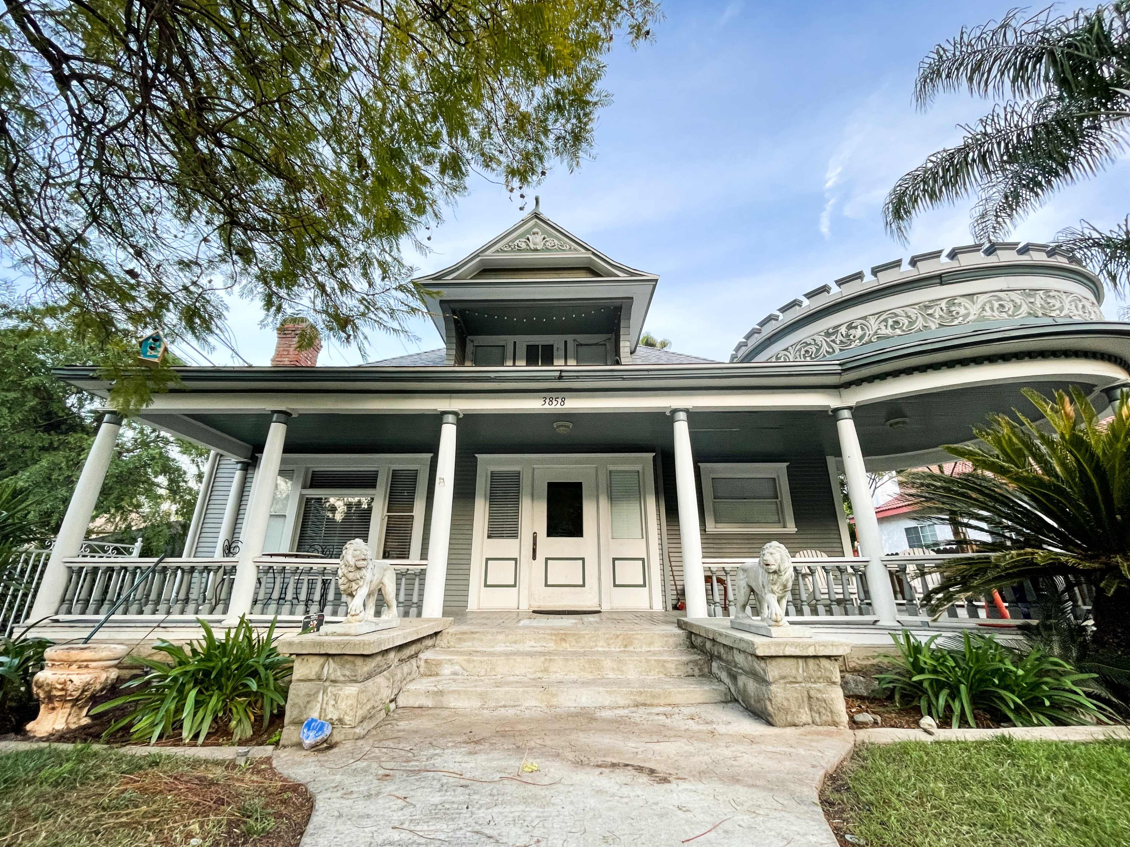 The image shows a large, historic house with a wraparound porch, ornate architectural details, and lush greenery in the front yard.