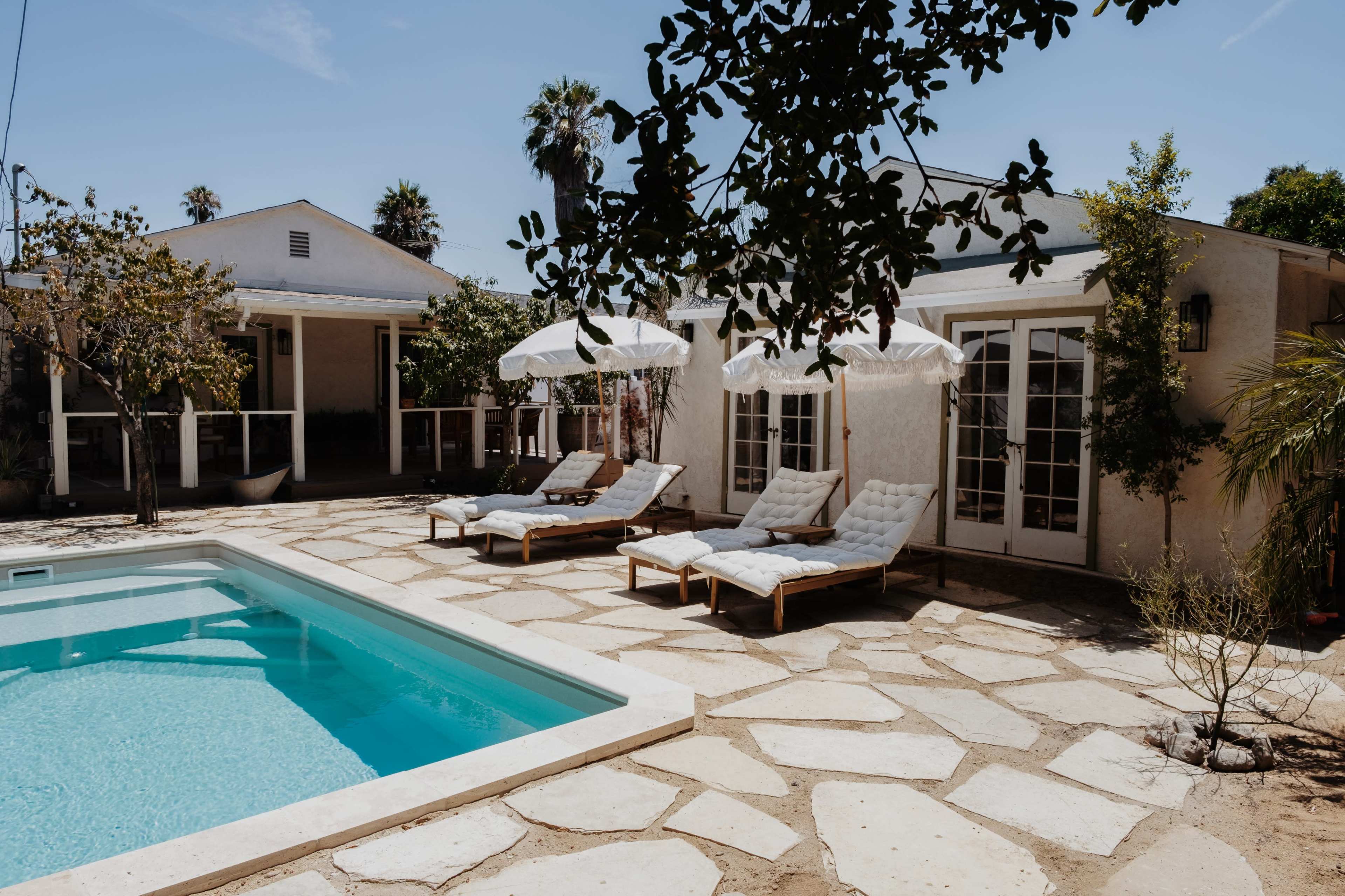 The image shows a pool area with two lounge chairs on a stone patio surrounded by palm trees and a house in the background.