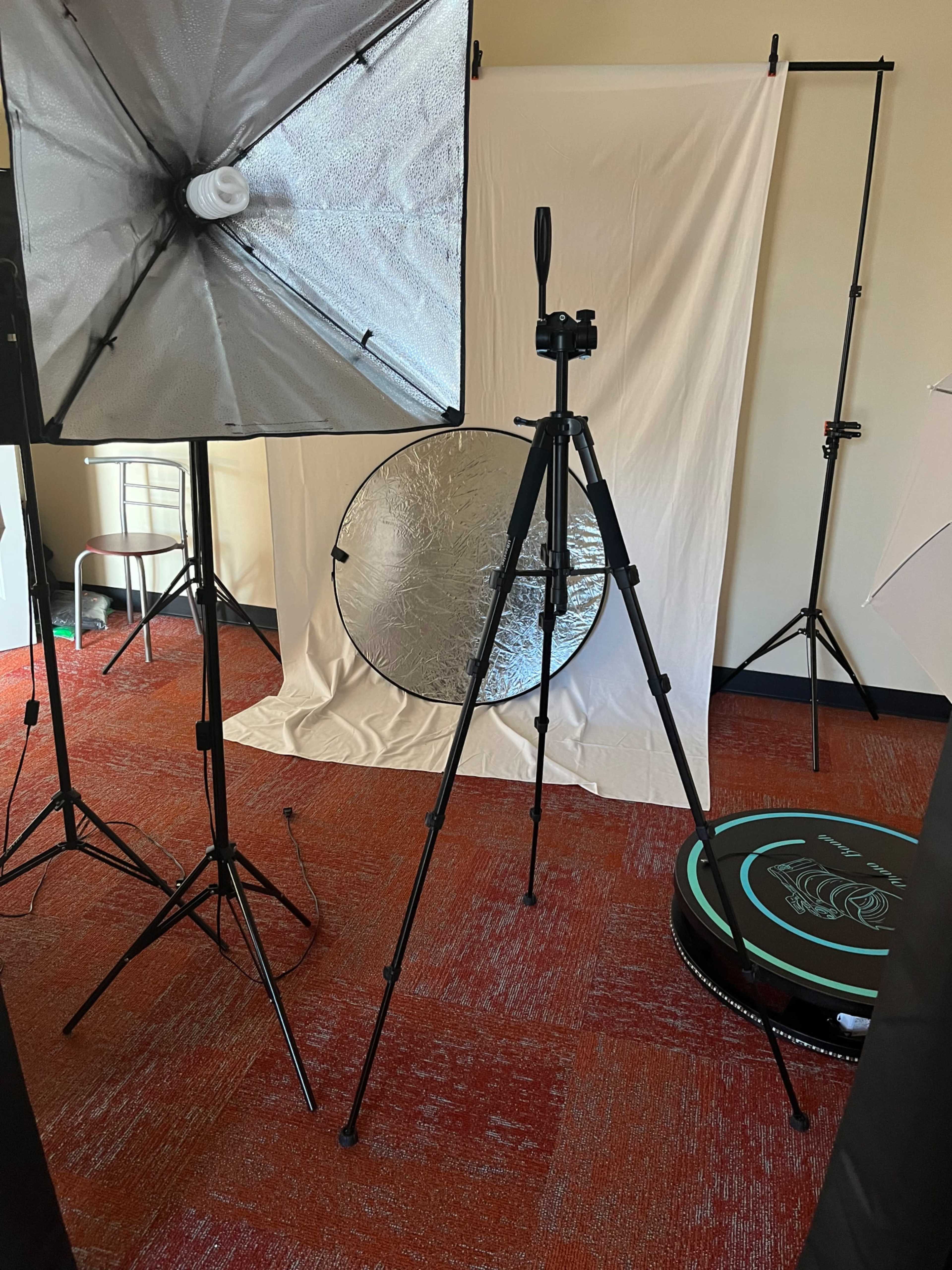 The image shows a photography setup featuring softbox lights, a camera on a tripod, a reflector, and a white backdrop in a studio environment.