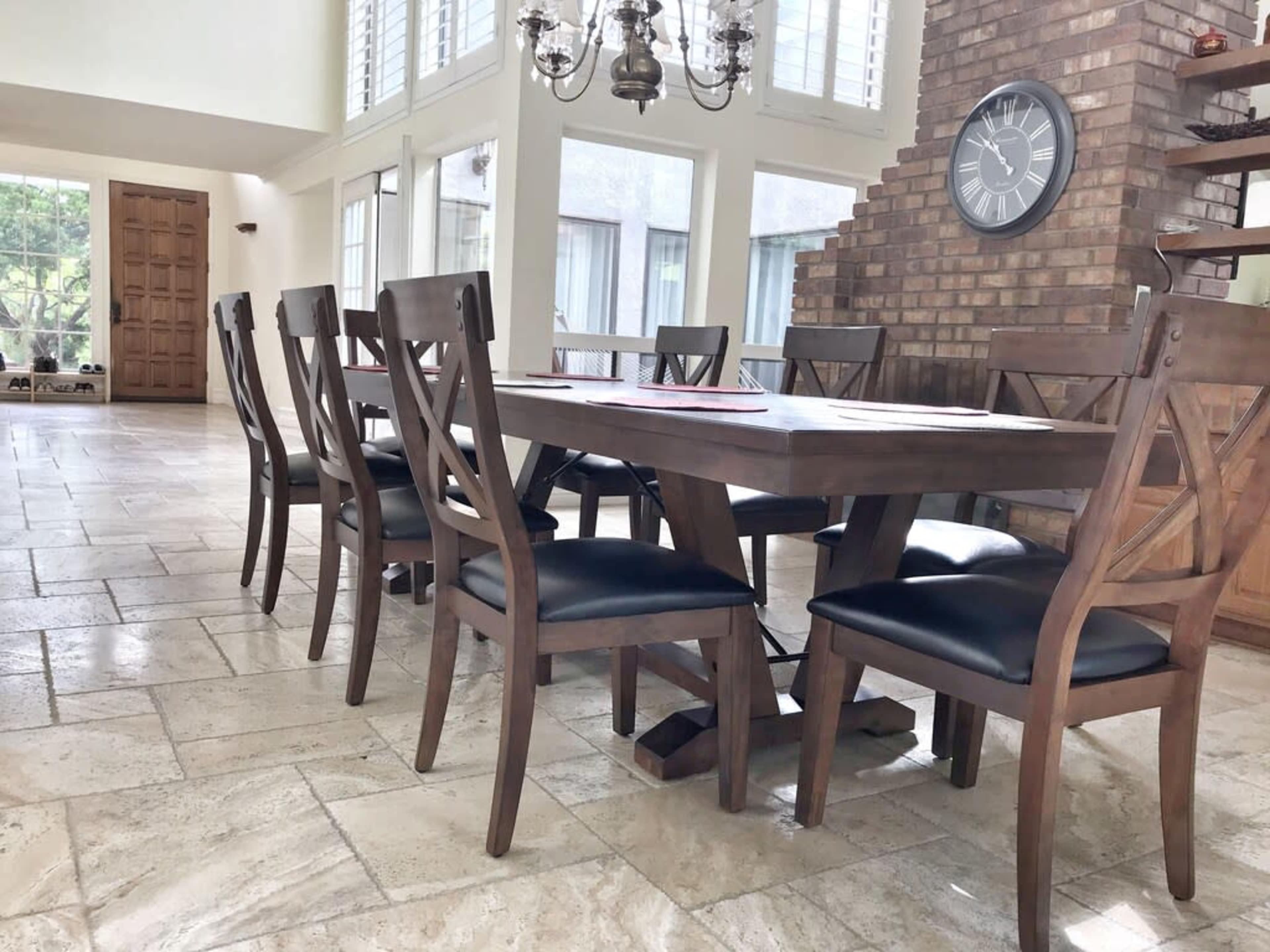 A wooden dining table with matching chairs is situated in a spacious room featuring large windows and a stone wall.