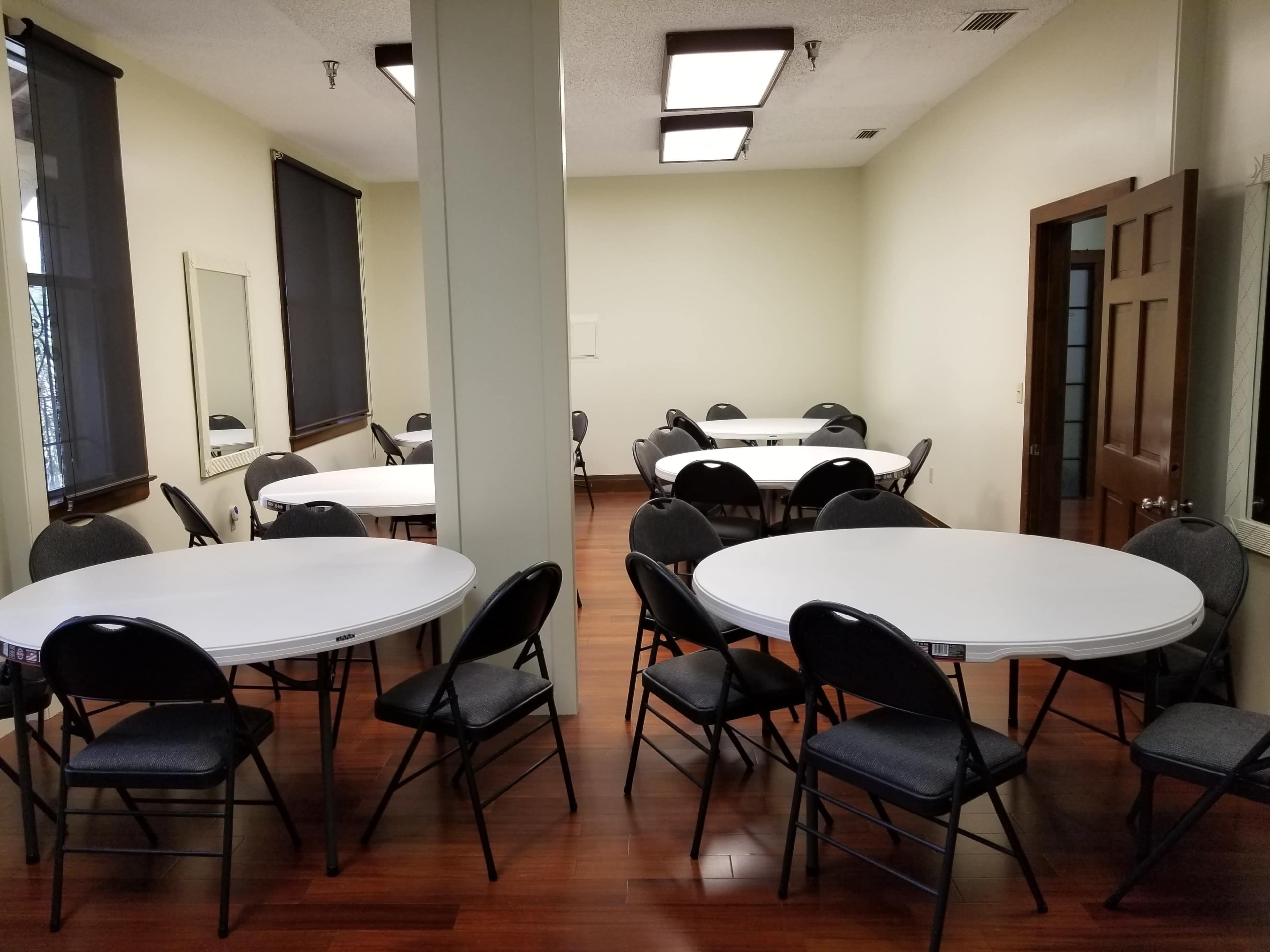 A room with multiple round tables and black chairs arranged around them, featuring hardwood flooring and large windows.