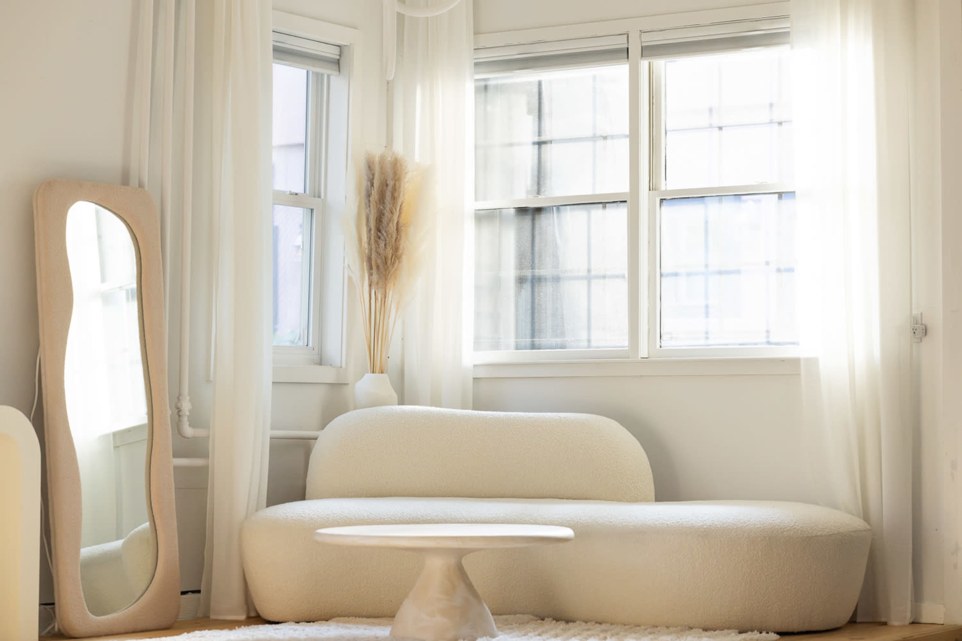 A light-filled room features a curved, cream-colored sofa, a round coffee table, and sheer curtains with a tall mirror beside a window.