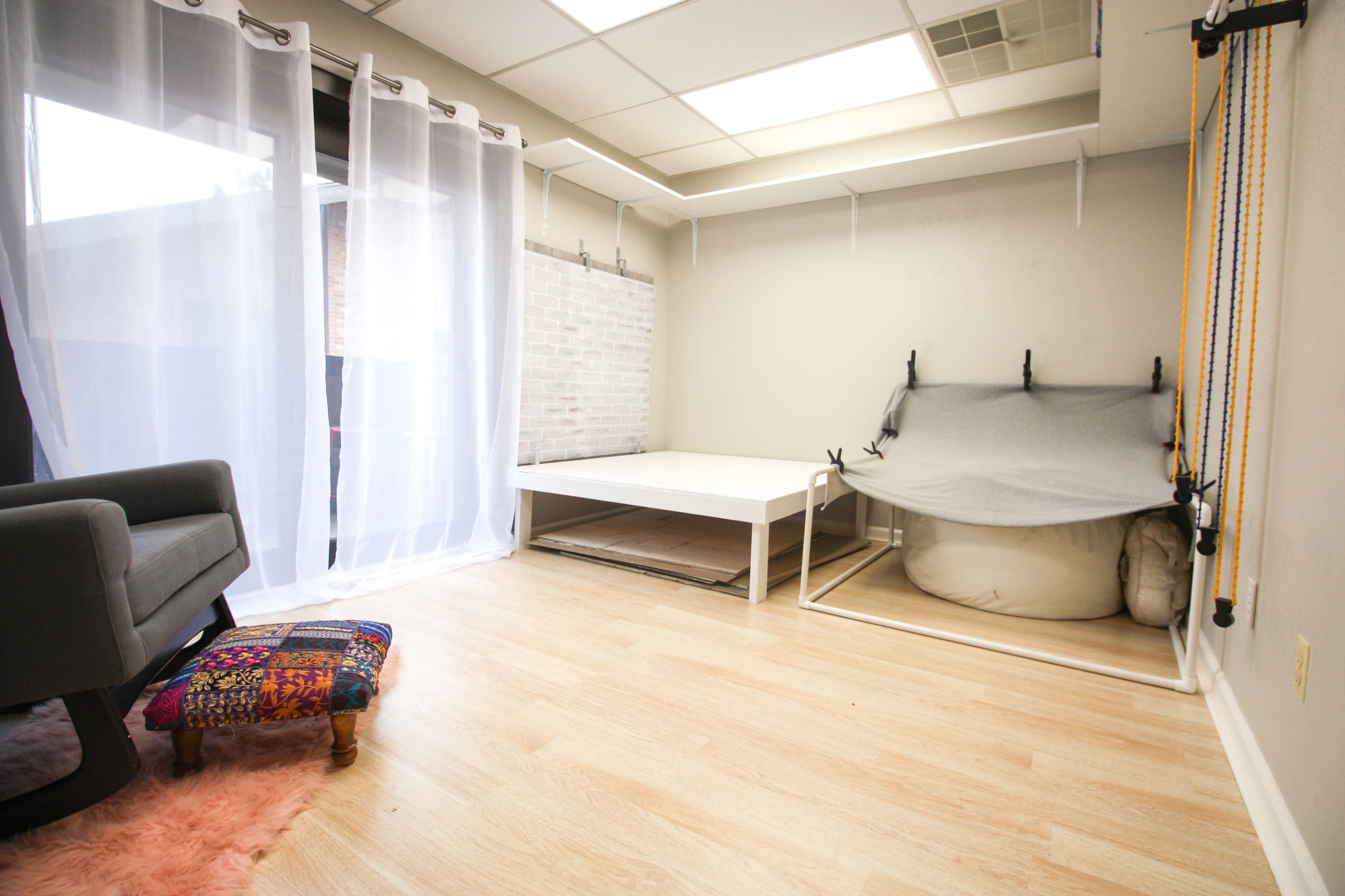 A minimalist room with light wooden flooring, a large window covered by sheer curtains, and a white platform bed alongside a hammock setup.
