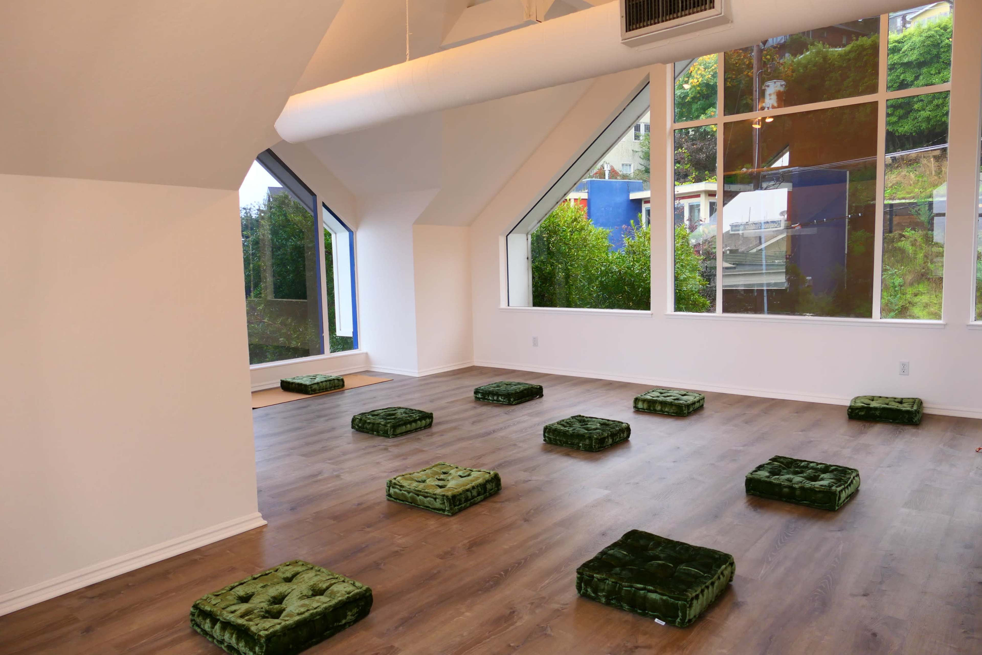 A minimalist room with large windows and green floor cushions spaced evenly on a wooden floor.