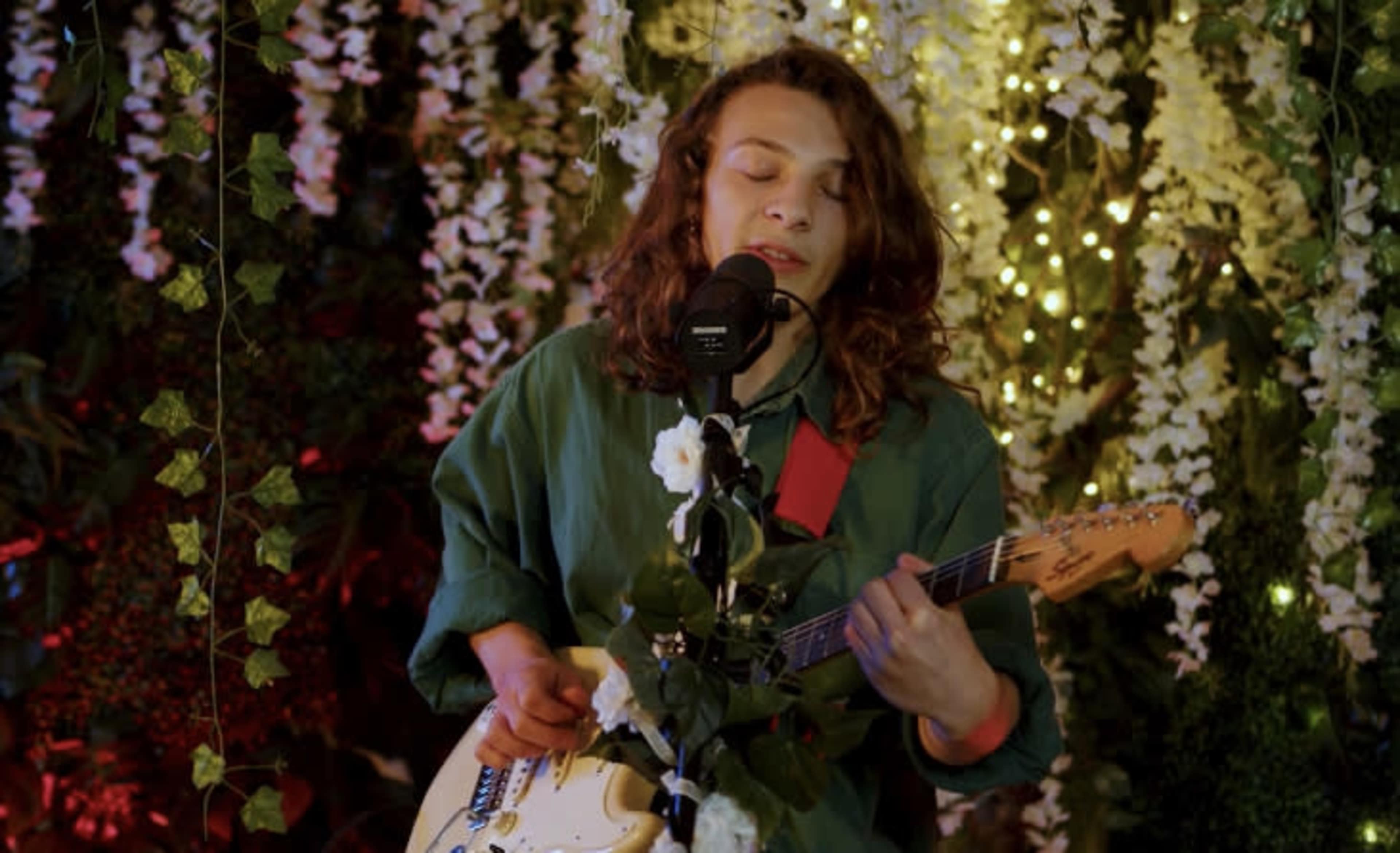 A musician plays an electric guitar while singing into a microphone adorned with flowers in a decorative setting filled with greenery and lights.