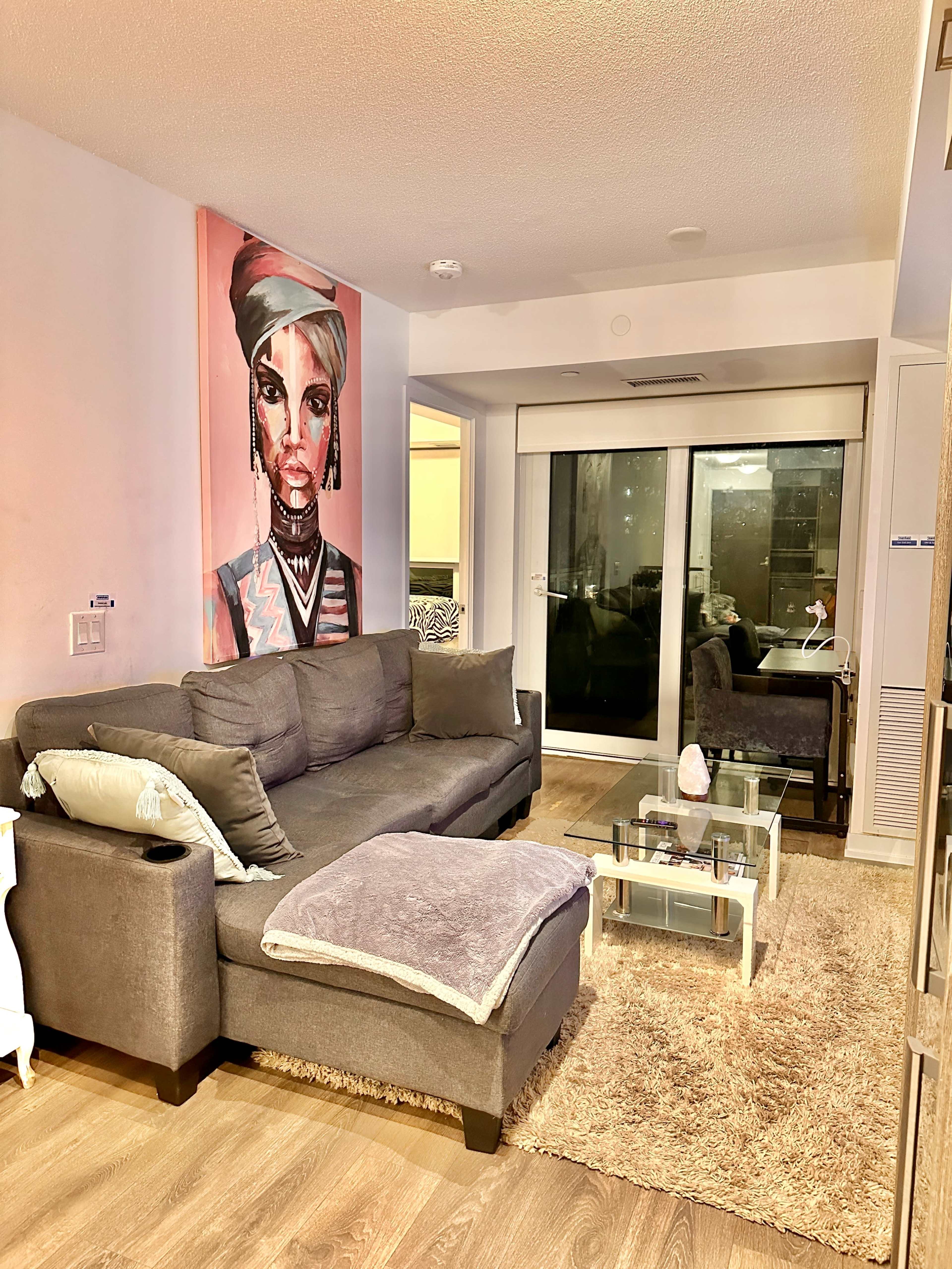 A modern living room features a gray sectional sofa, a glass coffee table, a large wall art piece of a woman, and a sliding glass door that leads to an outdoor area.